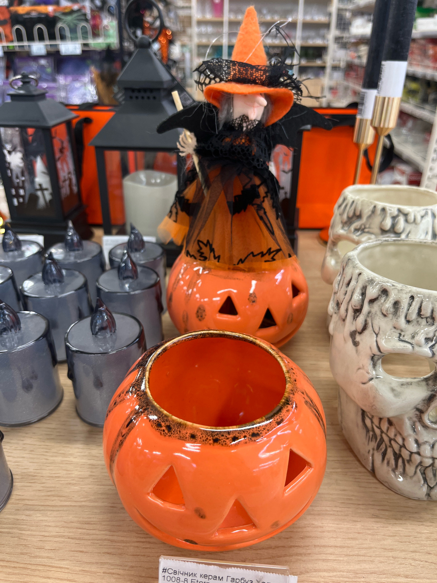 This is a close-up, eye-level, medium shot of Halloween-themed decorations displayed on a wooden surface, likely in a retail store. The focus is on an assortment of ceramic items. In the foreground, a bright orange, jack-o'-lantern shaped ceramic bowl with cut-out triangular eyes and a carved mouth is prominently featured. The bowl has black speckles near the rim and darker black stripes that mimic the natural ridges of a pumpkin. To the right of the jack-o'-lantern bowl, two white ceramic skull-shaped mugs are visible, with detailed carvings resembling dripping wax and skeletal features. Behind the jack-o'-lantern, a larger, more elaborate Halloween decoration is partially visible. This decoration appears to be a witch figure, complete with an orange pointed hat, black lace dress, and outstretched arms. The witch is perched atop another orange jack-o'-lantern, this one hollow and illuminated from within (though the light source is not directly visible). To the left of the witch figure, a row of grey, LED flameless candles is arranged. These candles have a matte finish and small dark gray tops resembling flames. Further in the background, two black decorative lanterns with cut-out designs suggesting spooky scenes are visible, placed on orange packaging material. The overall ambiance suggests a festive Halloween display. There is a small white tag with text partially visible at the bottom right, but the text is too blurry to be deciphered. The image is sharp and well-lit, with no people present in the frame. The retail environment is suggested by the blurred shelves and merchandise visible in the background. There is no specific information in the image to suggest a location in Kharkiv, Ukraine.