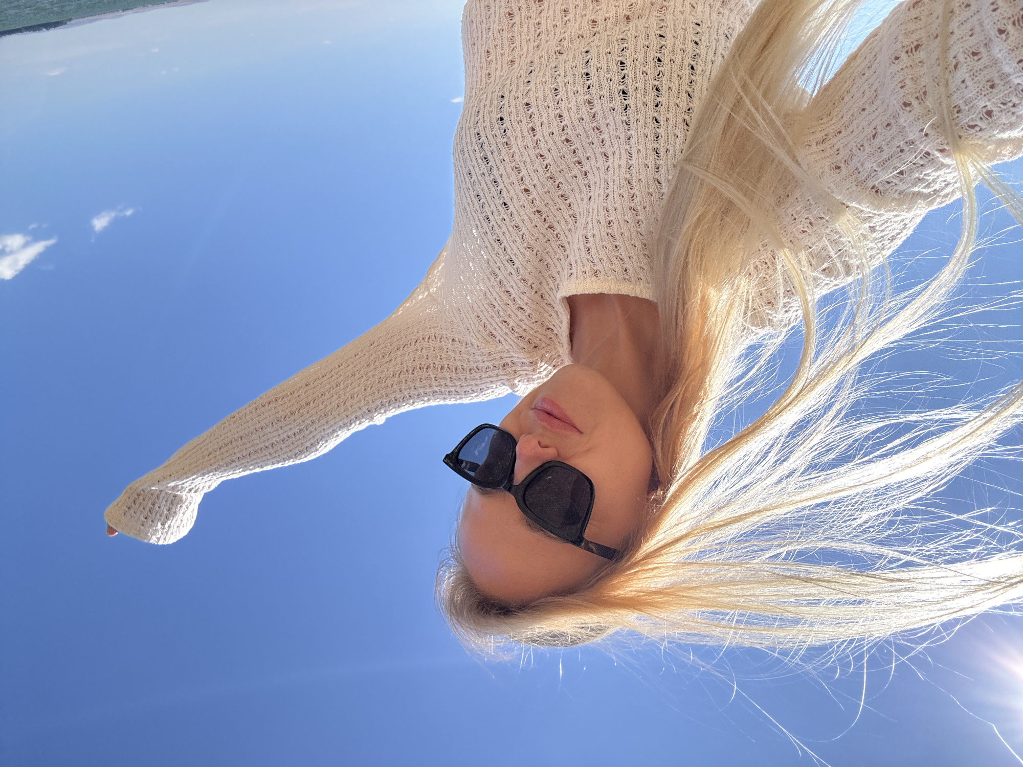 A blonde woman wearing sunglasses and a white, knitted sweater is taking a selfie outdoors under a clear blue sky. Her hair is blowing in the wind, and her right arm is raised. The sun is visible in the upper left corner of the image, creating a lens flare. The background appears to be a body of water with a distant shoreline. The location is identified as Gornyi Erik, Russia.