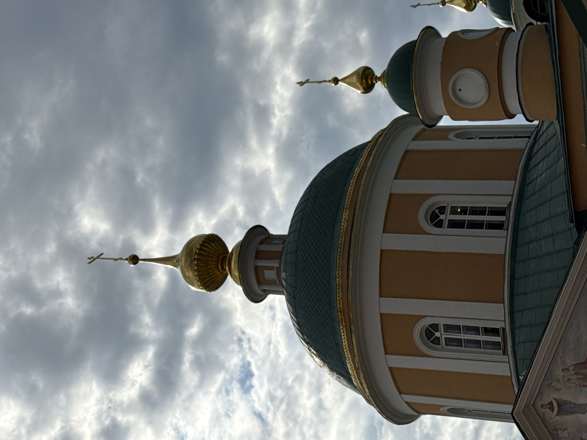A low-angle shot depicts a set of ornate church domes under a cloudy sky, possibly located in the city of Unknown, Unknown. The domes are a light tan color with white trim and are topped with green conical roofs and golden spires, each capped with a Christian cross. The sky is filled with grey and white clouds, indicating a partly cloudy day. The church's architecture, particularly the domes' design, suggests an Eastern Orthodox religious site.