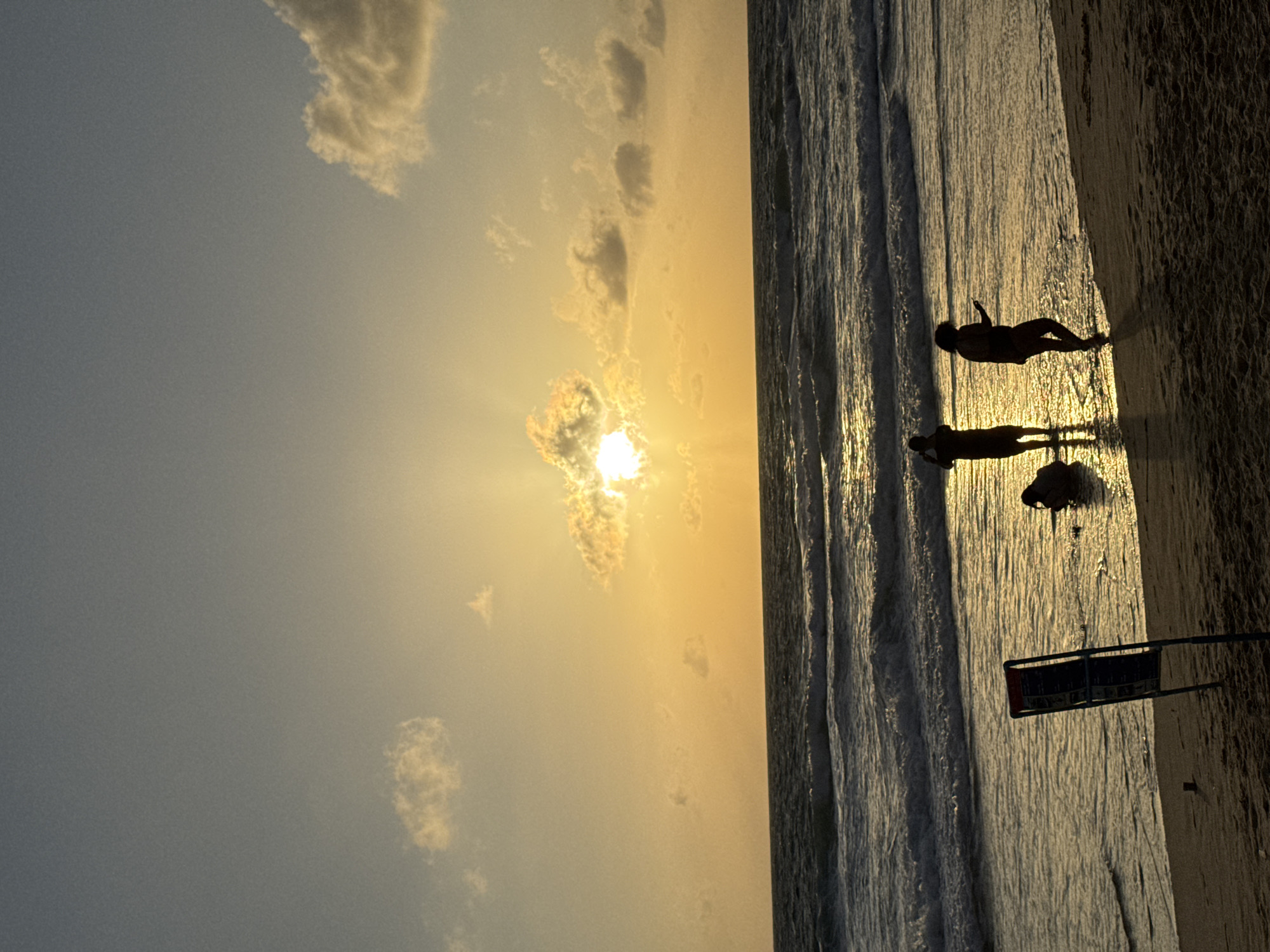 The image captures a serene sunset scene on Karon Beach in Karon, Thailand. The sun, a radiant golden orb, is partially obscured by a cloud, casting warm light across the sky and water. Gentle waves lap against the sandy shore where several silhouetted figures are present. One individual stands in the shallow water, looking towards the horizon. Another, possibly a child, is crouched down near the water's edge. Further down the beach, two other figures stand in the water, one holding a phone. A blue and red sign stands upright on the sand, near the water. The overall atmosphere is peaceful and tranquil, characteristic of a beach at dusk.