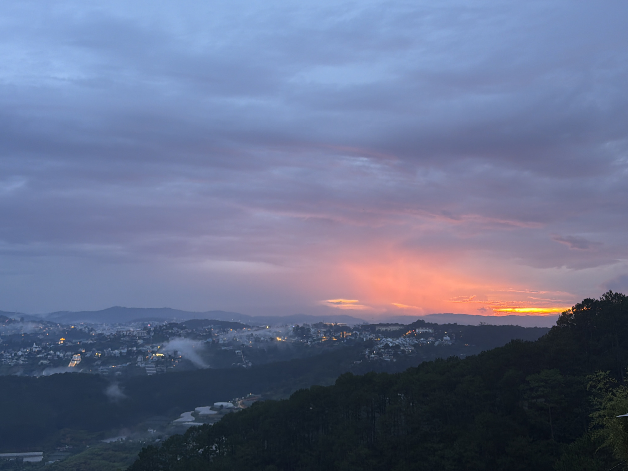 The image depicts a panoramic view of Xuan Truong - Da Lat, Vietnam, at dusk. A vibrant sunset paints the sky with hues of orange, pink, and purple, casting a warm glow over the landscape. Below, a sprawling city emerges from the mist, its lights twinkling like scattered stars. Buildings of various sizes and architectural styles dot the hillsides, interspersed with lush greenery. The foreground is dominated by a dense forest of pine trees, their dark silhouettes creating a dramatic contrast against the illuminated sky. The overall atmosphere is serene and picturesque, evoking a sense of tranquility and natural beauty.