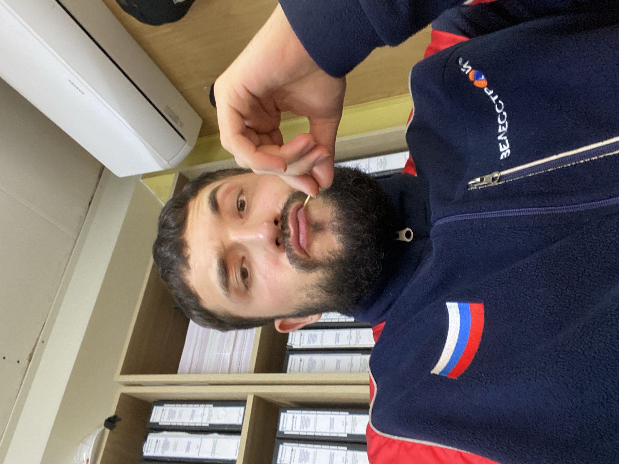 A man with a beard and dark hair is sitting in an office, holding a toothpick to his mouth. He is wearing a dark blue fleece jacket with red and white trim, and a Russian flag patch on the left chest. A logo with Russian text and colored circles is visible on the right chest. Behind him, shelves are filled with binders and files. An air conditioning unit is mounted on the wall above him. The scene appears to be in an office in Luzhitsy, Russia.