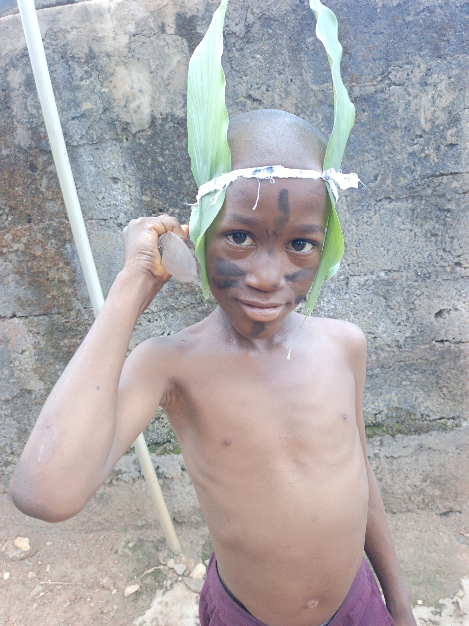 A medium shot captures a young, bare-chested boy of African descent, likely in Akwanga, Nigeria, posing outdoors against a weathered grey concrete block wall. He has a shaved head and wears a makeshift headdress made of large green leaves, secured by a white fabric strip tied around his forehead. His face is adorned with black paint, featuring a vertical stripe on his forehead and prominent smudges across his cheeks and nose.
The boy gazes directly at the viewer with bright, engaging eyes and a subtle, soft smile. He holds a small, light-colored, irregularly shaped object, possibly a stone or piece of wood, firmly in his raised right hand. A long, slender, light-colored pole or stick leans vertically near his left side, partially visible in the frame. He appears lean and is wearing dark purple shorts or trousers. The ground beneath him is uneven dirt mixed with small stones and debris. The bright, clear lighting suggests it is daytime. There is no visible text in the image.