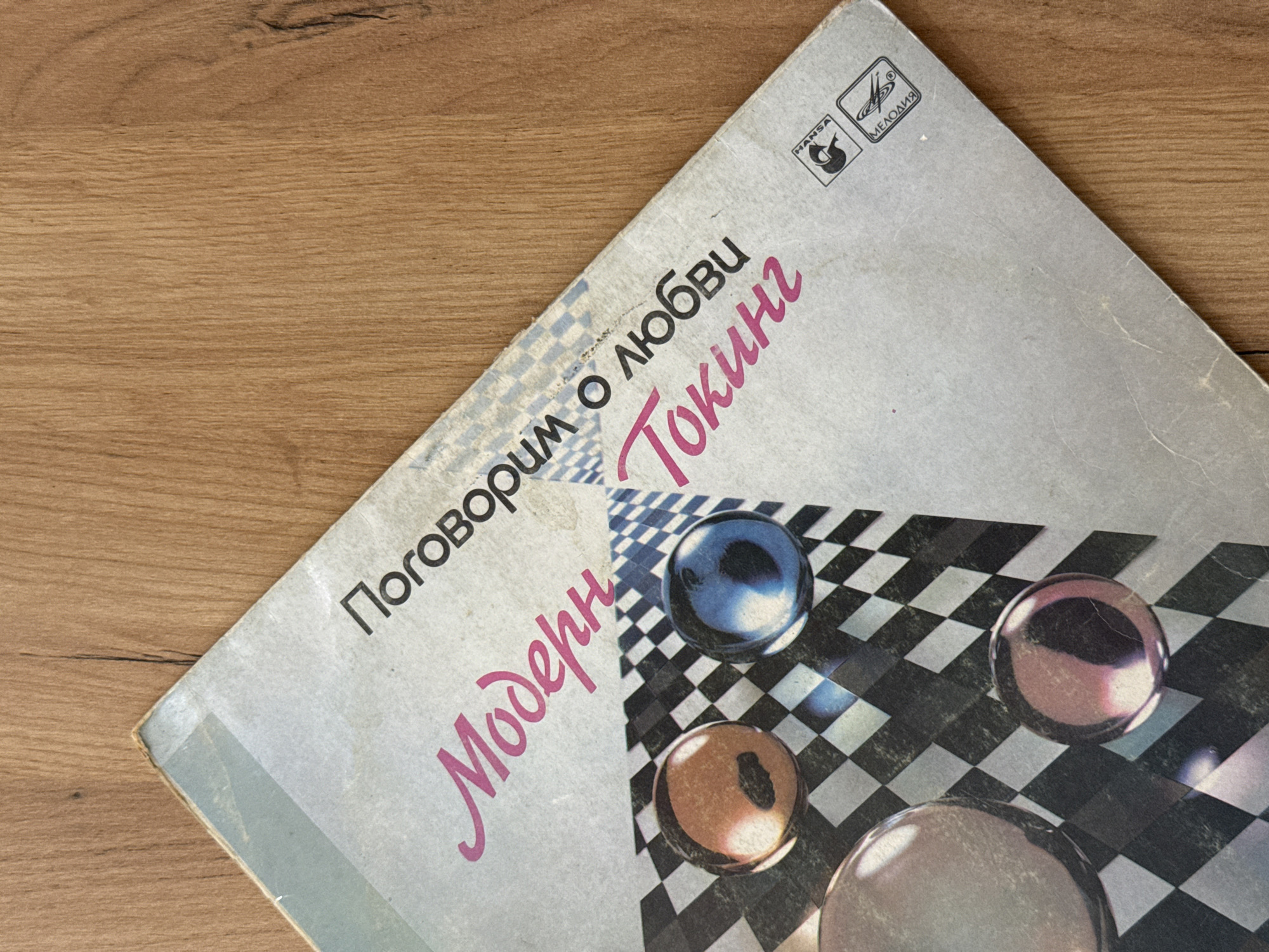 The image shows a close-up, overhead view of an old record album cover lying on a wooden surface. The album cover is tilted diagonally from the top left to the bottom right corner. The cover is primarily light grey with pink and black text. The pink text, in a stylized script, reads "Модерн" (Modern). The black text, in a sans-serif font, reads "Поговорим о любви" (Let's talk about love). Below this, in pink, is the word "Токинг" (Talking). The background of the album cover features a black and white checkered pattern with some abstract blue and white geometric shapes. Three glass marbles are visible on the checkered pattern: one is orange with a black shadow, another is blue with a black shadow, and a third, larger marble is at the bottom left, appearing to reflect the checkered pattern and pink text. In the bottom right corner of the album cover are two logos. The top logo is a black silhouette inside a square with the word "HANSA" above it. The bottom logo is a stylized black and white design within a square with the word "МЕЛОДИЯ" below it. The wooden surface appears to be a tabletop with a visible wood grain pattern. There are no people or any identifiable location in the city of Nizhny Novgorod, Russia depicted in this image.