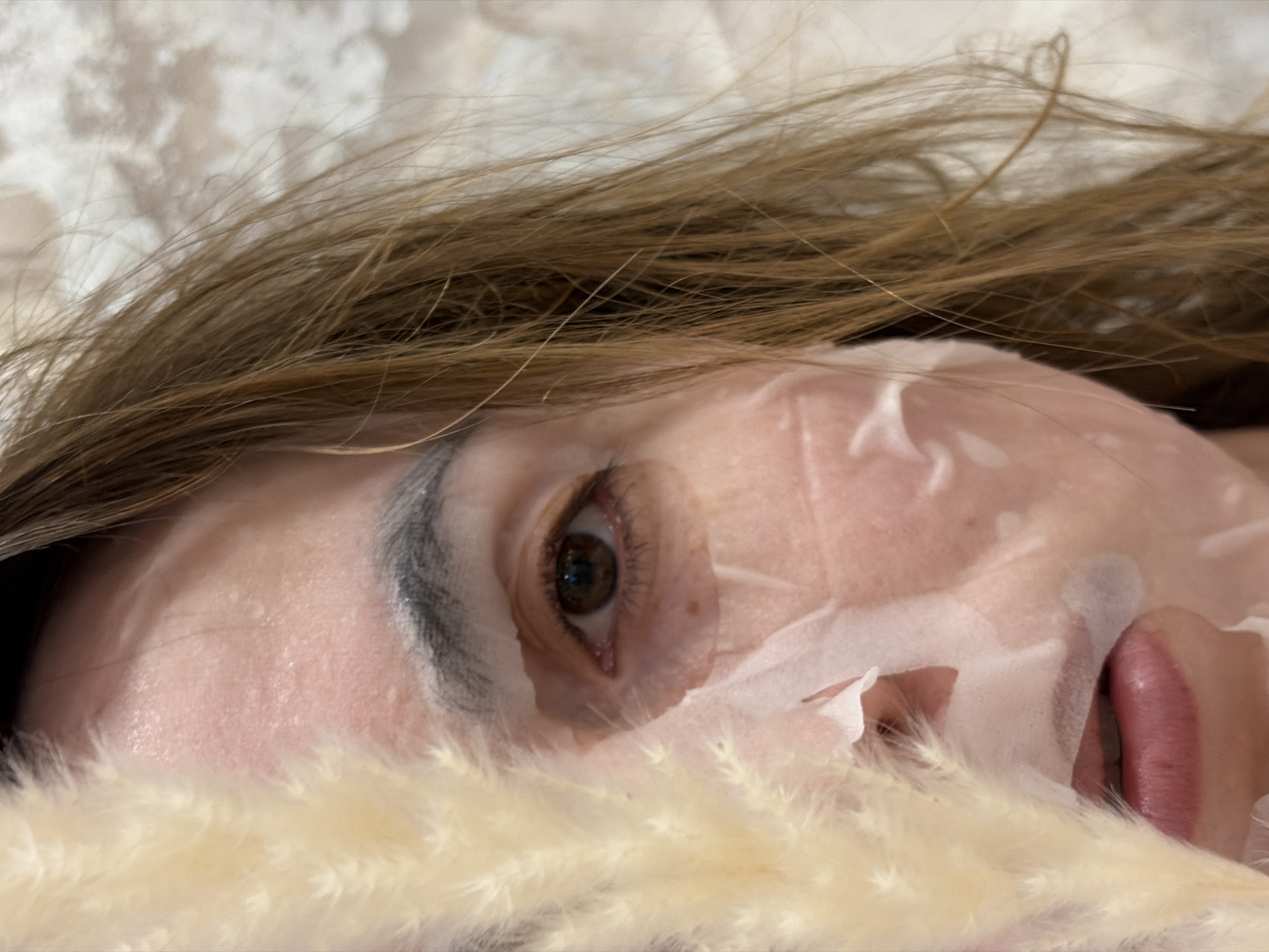 A close-up shot reveals a woman's face covered in a sheet mask, seemingly lying down with her head slightly tilted. The mask has some creases and tears, particularly around the nose area. Only one eye is visible, its color appears to be brown. She has brown hair. A soft, fluffy decorative element, possibly pampas grass, is partially obscuring her lower face.
There is no information in the image to determine the location. Therefore, it is impossible to provide any information about Izhevsk, Russia.