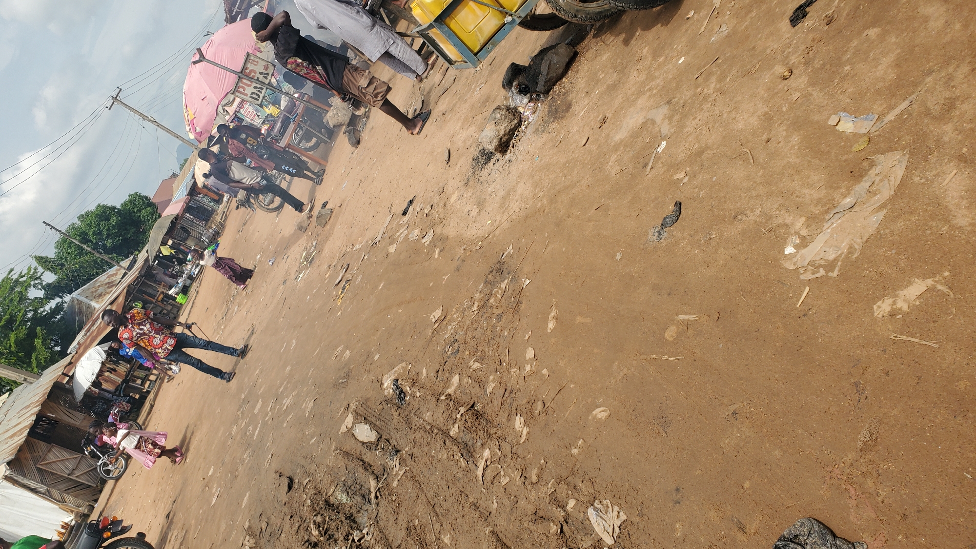 The image depicts an outdoor scene in Jalingo, Nigeria, likely a market or roadside area. The ground is composed of dirt and mud, with scattered debris, including plastic sheeting and sticks. Several individuals are present, dressed in casual clothing.
In the foreground and midground, a man wearing a brightly colored, patterned shirt and dark pants stands with a stick or pole in his hand. To his left, a young girl in a pink dress is captured mid-movement. Further back, several other people are visible, some standing near what appear to be market stalls or shelters with corrugated metal roofs. Motorcycles are parked in various locations. A large pink umbrella is prominent in the background, providing shade over a stall. In the right side of the image, a yellow container on a wheeled cart is visible. The overall environment appears to be somewhat unkempt and active with daily life.