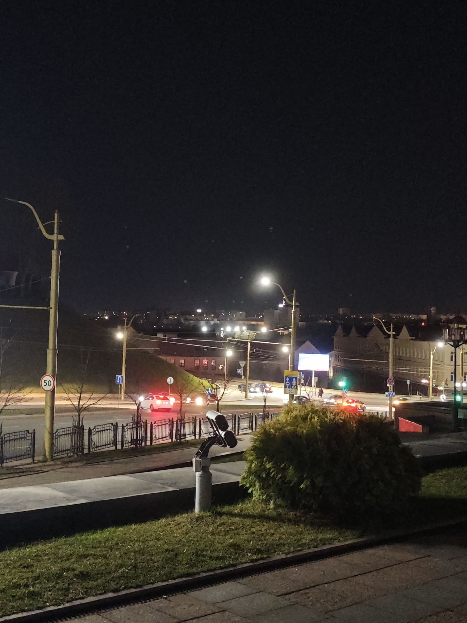 The image depicts a nighttime urban scene in Hrodna, Belarus, observed from an elevated position, likely the Old Bridge (Stary Most), looking towards the intersection of vulitsa Dzerzhinskogo and vulitsa Mostovaya, with vulitsa Karla Marksa visible in the background. The environment is illuminated by artificial light sources against a dark sky.
In the immediate foreground, a paved surface transitions to a strip of green grass. A grey cylindrical pole with a black multi-camera or sensor apparatus mounted at an angle directs its view towards the street below. A dark metal fence with ornamental scrollwork separates this elevated area from the roadway.
The midground features a multi-lane asphalt road, appearing wet or reflective. Several vehicles are present: a white sedan with illuminated red taillights proceeds right in the left lane, followed by another vehicle. At the intersection to the right, multiple vehicles exhibit illuminated red taillights, with some headlights visible. One vehicle appears to be a taxi with an illuminated roof sign. A green traffic signal is active on the right. Streetlights on poles cast light on the road. A circular regulatory sign displaying "50" within a red border indicates a speed limit. A blue square sign with a white U-turn arrow is also visible. Bare trees line a grassy, ascending embankment on the left side of the road. A large, dense green bush partially obscures the view on the mid-right.
The background reveals urban structures, including a prominent older building with a distinctive multiple-gable roofline on the right side of the street (vulitsa Karla Marksa). A brightly illuminated rectangular billboard or digital display is positioned near the intersection. Distant city lights punctuate the dark horizon. One pedestrian is faintly discernible near the intersection. No specific interactions between individuals or vehicles are clearly identifiable.