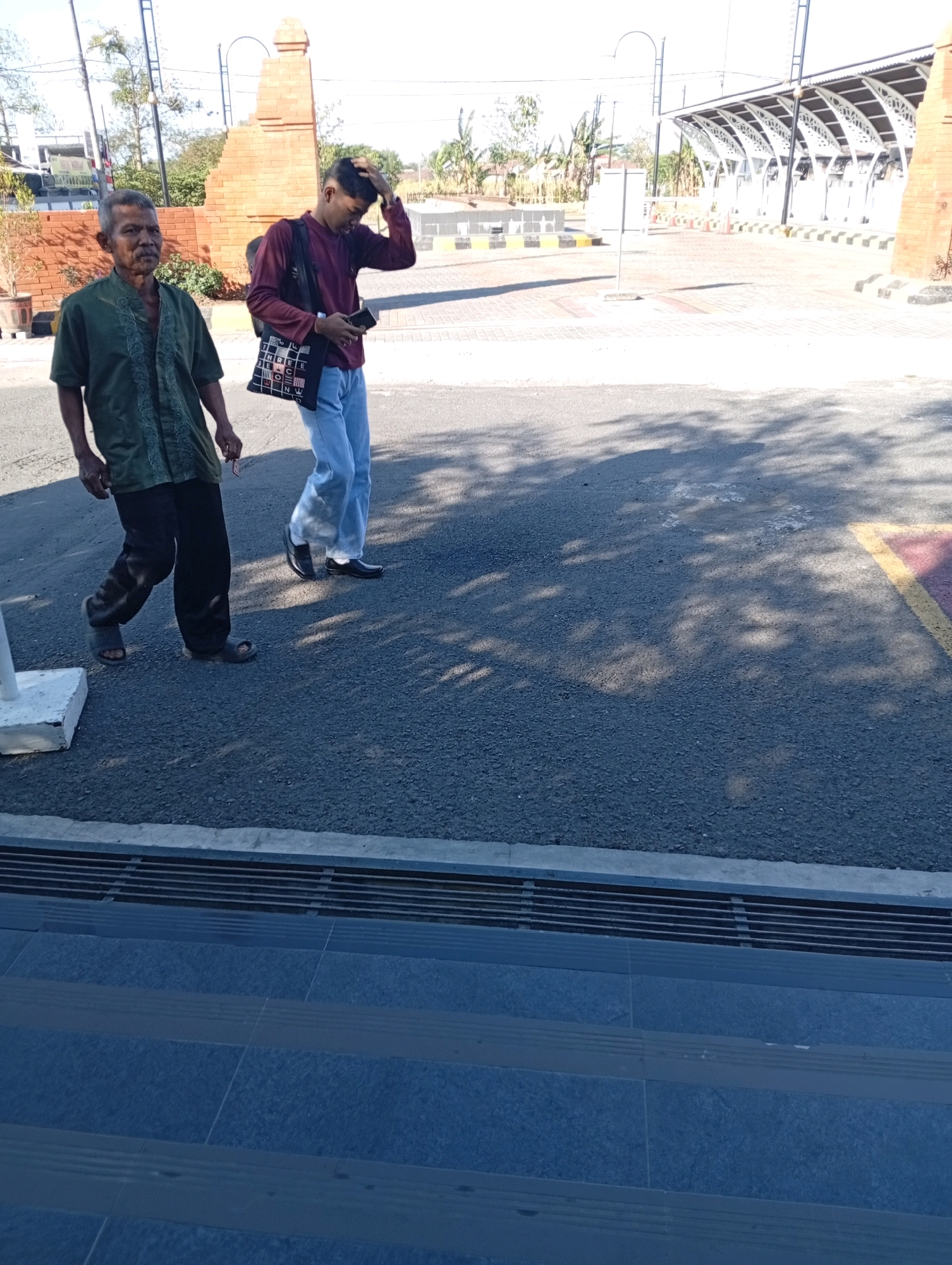 Two men are walking on a paved area outdoors. The man on the left is older, wearing a green, collared shirt and dark pants with sandals. He is looking towards the camera. The man on the right is younger and wearing a maroon long-sleeved shirt, light blue pants, and black shoes. He is holding a black smartphone in his right hand and has his left hand raised to his head. He is looking down at his phone. Behind the men are brick structures and a covered walkway with a white roof. The ground is asphalt with some shadows cast on it. The scene appears to be in a public area, possibly a transportation hub or a park.