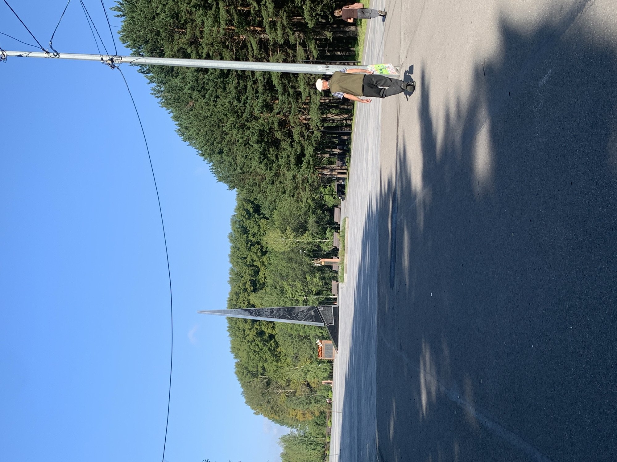 The image depicts an outdoor scene in the city of Prokopyevsk, Russia, specifically within a memorial park. In the foreground, a paved walkway or road is visible, with shadows of trees cast upon it. Two individuals are walking away from the camera towards the right. The person in the foreground is an elderly man wearing a white cap, a plaid shirt, and a dark vest, carrying a multi-colored shopping bag. Behind him, a woman in a brown top and dark pants is also walking.
In the mid-ground, a large, abstract memorial sculpture dominates the view. It is a tall, dark, triangular structure adorned with relief carvings or etchings. To the left of the main sculpture, there is a smaller monument with a red ribbon-like element. The memorial area is paved with light-colored stones and surrounded by numerous park benches.
The background features a dense line of tall pine trees, suggesting a wooded area or a park setting. The sky above is clear and blue. A utility pole with wires runs along the right side of the image. The overall impression is that of a quiet, sunny day in a public space dedicated to remembrance.