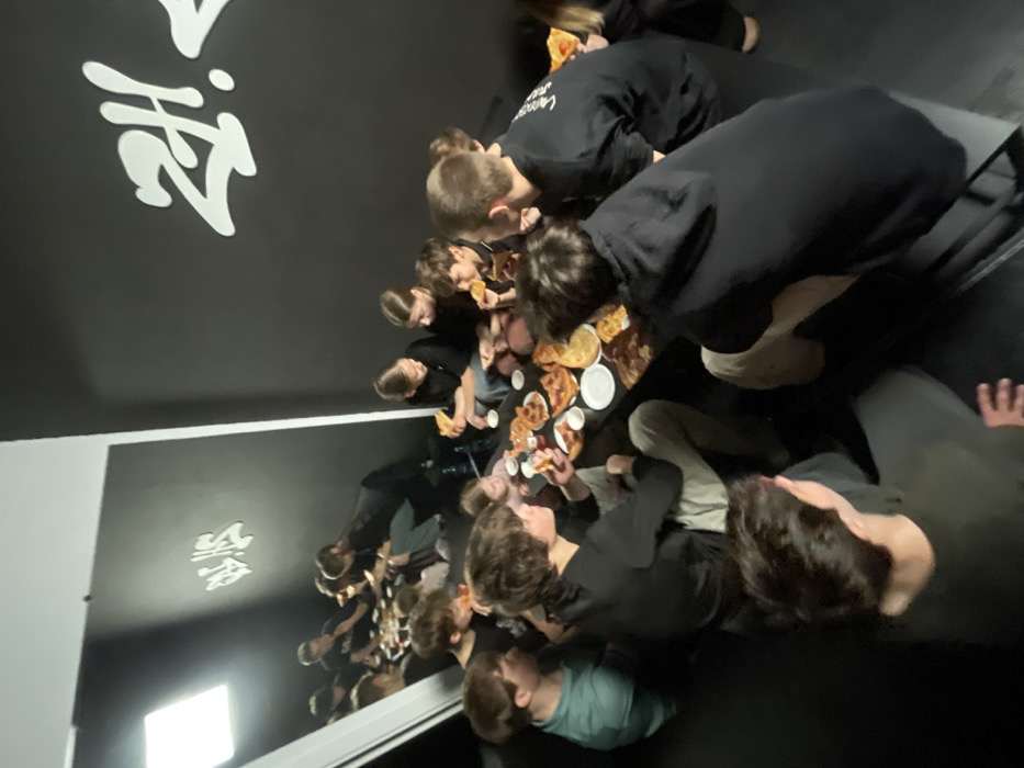 The scene depicts an interior room, likely a contemporary dance or youth studio, located at Mechnikova Street, 45, Krasnoyarsk, Russia. The room features matte black walls; one prominently displays a large white graffiti-style "ЖИ!" (ZHI!) logo. A significant portion of the left wall is covered by a mirror, reflecting the room, its occupants, and a bright, square overhead light source. Approximately fifteen individuals, predominantly teenagers and young adults, including one younger girl, are gathered around a low, rectangular dark table. They are dressed casually, primarily in dark clothing. The group is actively consuming pizza; multiple pizzas, individual slices on white paper plates, and condiment bottles (including red sauce) are dispersed across the table. Beverages in plastic bottles are also visible. Individuals are observed holding and eating pizza slices, engaging in social interaction within the informal setting.