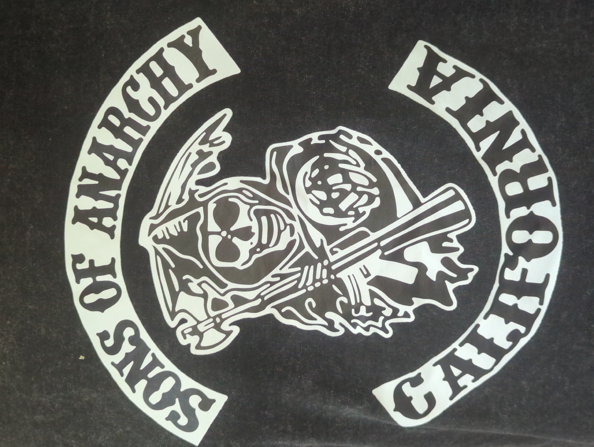 The image shows a close-up of a dark grey t-shirt with a white graphic. The graphic features a skull wearing a helmet and holding a scythe, with the words "SONS OF ANARCHY" arched above it and "CALIFORNIA" arched below it. The t-shirt is likely a piece of apparel related to the television series "Sons of Anarchy". There is no indication of the city of Iserlohn, Germany, in the image or its contents.