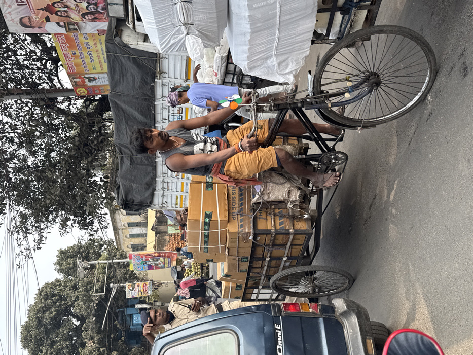 A man pedals a heavily loaded cycle rickshaw. Boxes strapped together with rope dominate the rickshaw's cargo space. The man wears a tank top and shorts. Another man walks alongside the rickshaw. A police car with an officer visible inside is parked behind the cycle rickshaw. A building with an arch and vendors selling fruit can be seen in the background. The overall scene depicts everyday commerce in a bustling area, possibly in India, judging by the signage in the background.