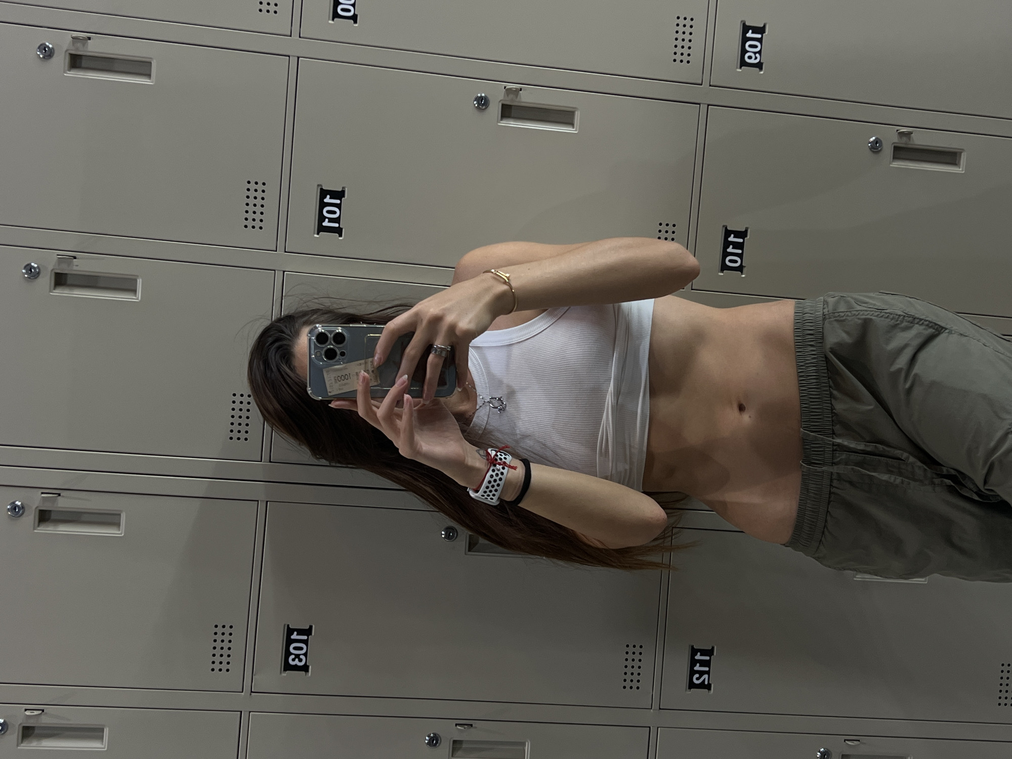 A person stands in front of a bank of lockers, holding a smartphone to take a mirror selfie. The person is wearing a white tank top and gray cargo pants. The lockers are beige with black numbers on them, and the background is a neutral gray color. The location is Unknown, Unknown.