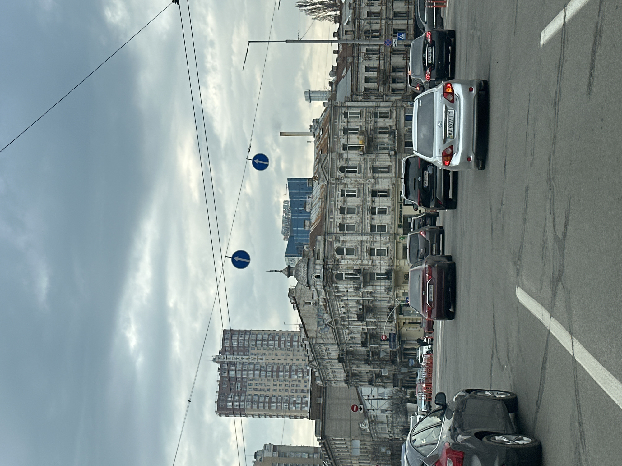 The image depicts a street scene in Kyiv, Ukraine. Cars are lined up in traffic on a city street. Buildings are visible on both sides of the road, with a mix of older, ornate structures and more modern high-rise buildings. Traffic signs indicating directions are present. Overhead wires stretch across the sky. The sky is overcast, suggesting a cloudy day.