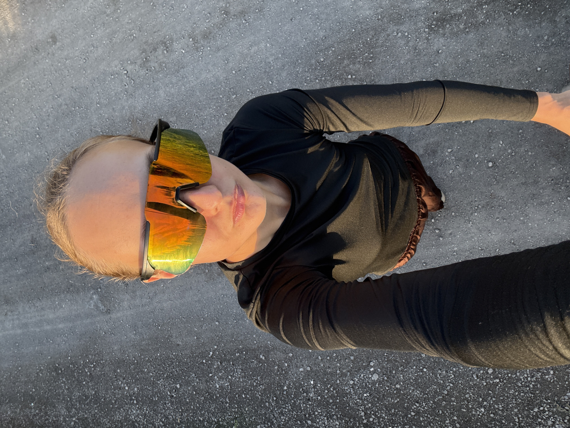 A person stands on a gravel surface, facing upwards towards the camera. They are wearing a long-sleeved black shirt, brown shorts, and large, mirrored sunglasses with orange and yellow reflections. The person's blonde hair is visible at the top of their head, and their face is partially obscured by the sunglasses. The background consists of the rough, gray gravel, with some sparse white pebbles. The scene appears to be outdoors, with sunlight casting shadows on the person and the ground. There are no buildings or other identifiable structures visible in the image. The exact location within Podshivalovo, Russia, cannot be determined from the visual information provided in the image.