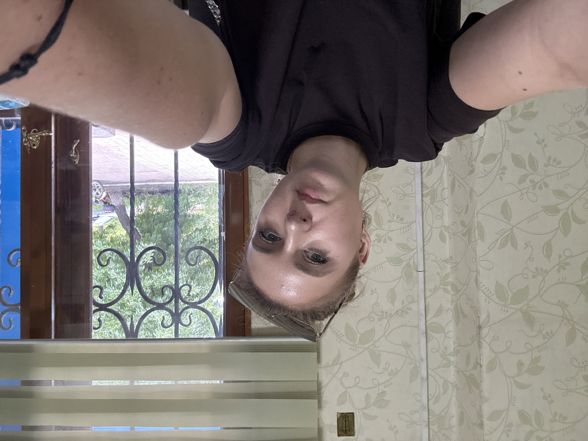 A person is taking a selfie in what appears to be a room with patterned wallpaper. The person is wearing a black t-shirt and has sunglasses perched on their head. Their face is turned towards the camera, with visible makeup on their eyes and lips. In the background, there is a window with a view of greenery and some decorative metal bars. A set of blinds is visible below the window. The lighting suggests it could be daytime. The image is oriented upside down. There is no visible text.