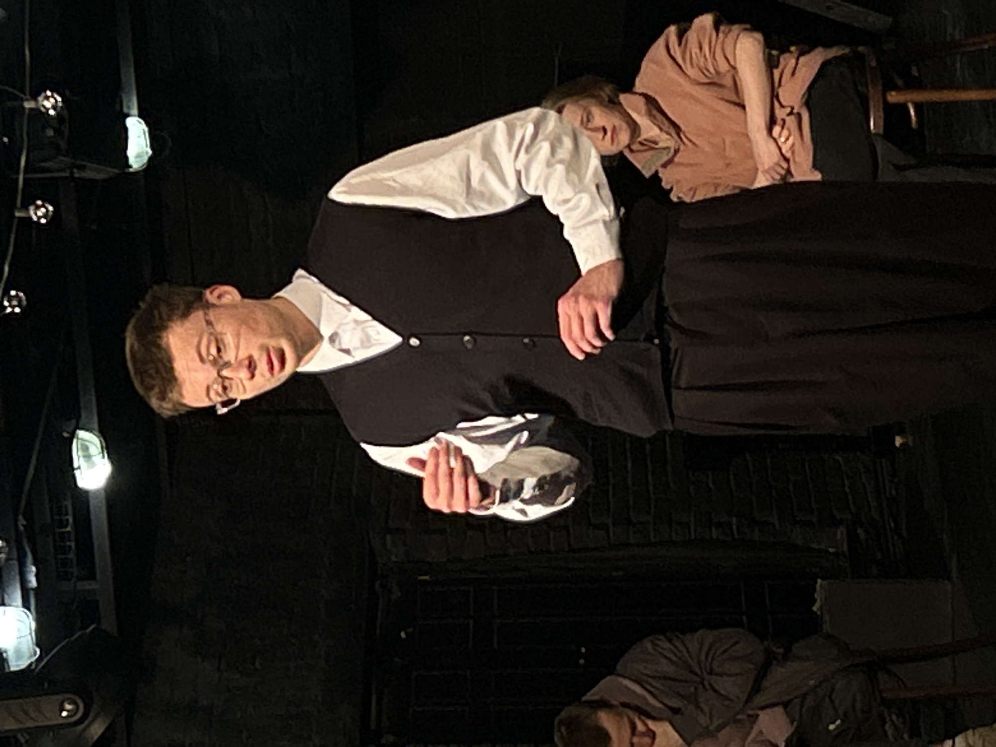 The image, rotated 90 degrees counter-clockwise, depicts an indoor theatrical scene, likely a performance or rehearsal.
In the center-left, a man with light brown hair and glasses stands, wearing a white collared shirt, a dark vest, and dark trousers. He appears to be actively performing, with his mouth open as if speaking or singing, and his hands clasped in front of him. He looks directly towards the viewer.
To the upper right, another person with lighter hair, dressed in a loose-fitting, long-sleeved peach-colored shirt and dark trousers, leans against a dark background. Their head is rested, and their eyes seem closed or cast downwards, suggesting a contemplative or resting state. Part of a wooden chair back is visible beside them.
In the bottom right corner, a third person is partially visible, lying or crouching on a dark surface, wearing dark clothing. Only their head and upper torso are discernible.
The background consists of dark, textured walls, possibly stage flats or brickwork. Several bright stage lights are visible along the left edge of the frame, casting a dramatic illumination on the central figure while the rest of the scene remains relatively dim. The overall lighting and setting strongly indicate a staged event in a performance space. No visible text is present.