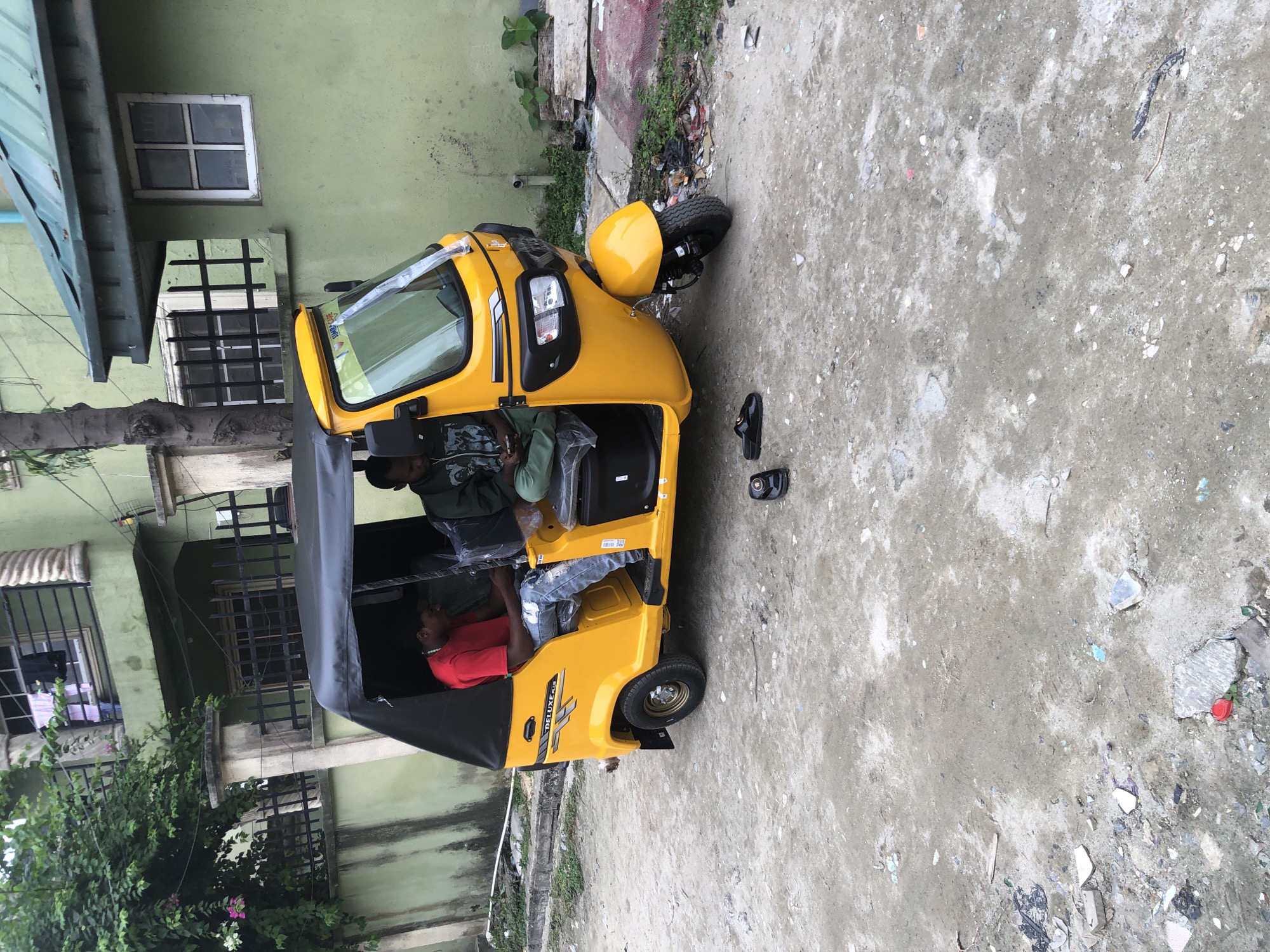 A yellow three-wheeled auto rickshaw, identified by the "DELUXE PLUS" branding on its side, is parked on a dirt and debris-strewn street in Ado, Nigeria. Two men are seated inside the vehicle. The driver, on the left, wears a red t-shirt and ripped blue jeans. The passenger, on the right, is dressed in a green hoodie and light green pants. A pair of black slip-on sandals are placed on the ground near the passenger side of the auto rickshaw. The background features a two-story building painted light green, with barred windows on the lower level and a balcony on the upper level. Lush green trees and foliage are visible to the left of the building. The overall scene depicts a typical street view in a Nigerian urban environment.