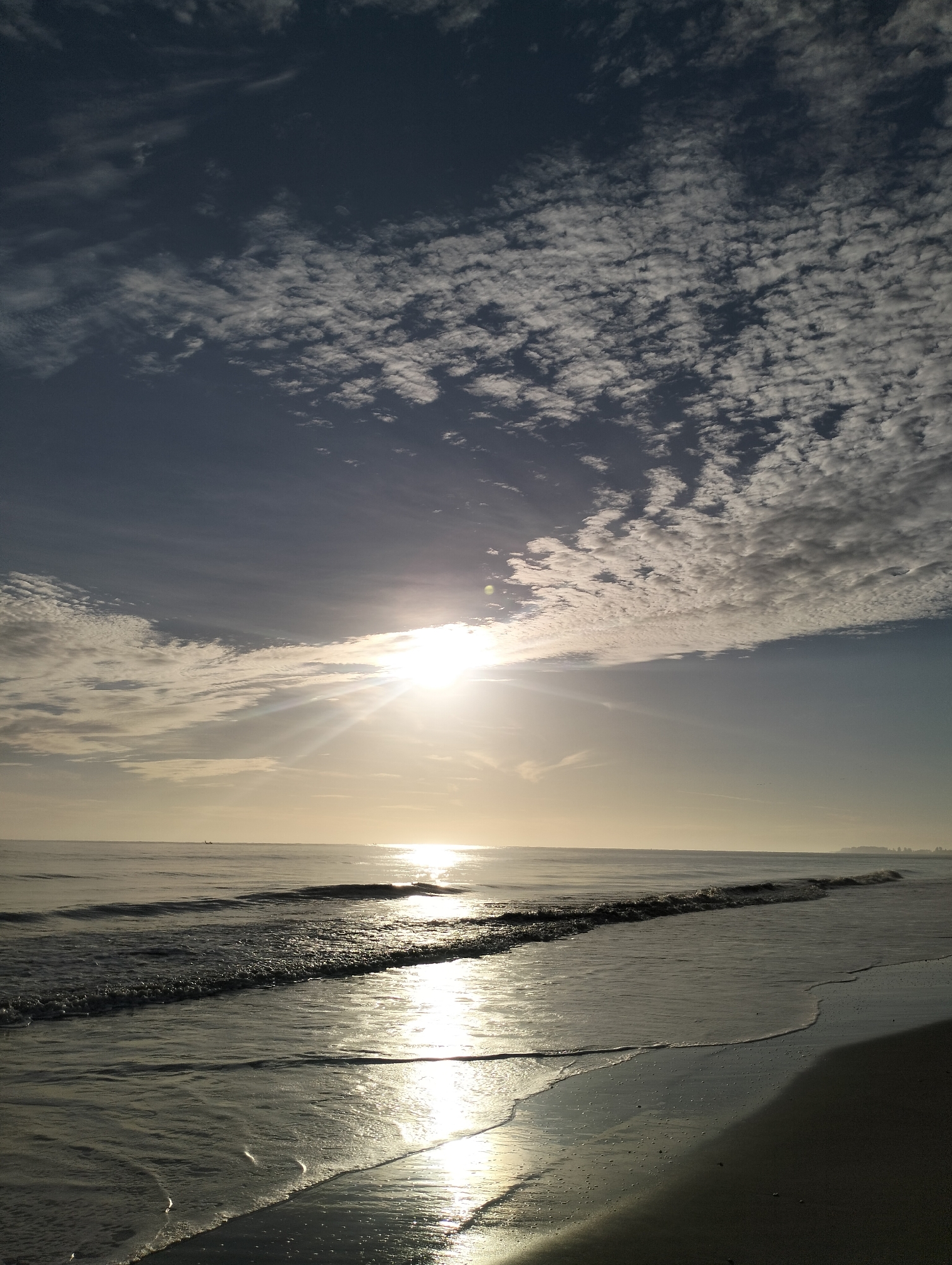 The image depicts a beach scene with the sun shining brightly above the ocean. The sky is filled with scattered clouds. The sunlight reflects off the water, creating a glistening effect. The shoreline is visible in the foreground, with gentle waves lapping onto the sand. The beach is located in the city of Sagoe, Indonesia.