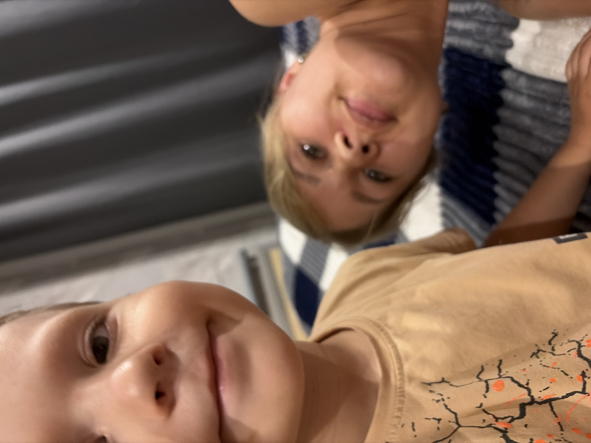 In the foreground, a young child's face is captured from a close-up perspective, their mouth slightly open as if speaking or laughing. They are wearing a light brown t-shirt with a graphic print. To the right, further in the background and out of focus, a young woman is lying down, her head tilted. She has blonde hair and is wearing a small earring. The background consists of a draped grey curtain and a blurred plaid blanket with blue and white stripes. The image appears to be taken indoors, possibly in a bedroom, with the lighting suggesting it might be evening or a dimly lit room. There are no obvious landmarks to suggest a specific location within Chaikovsky, Russia.