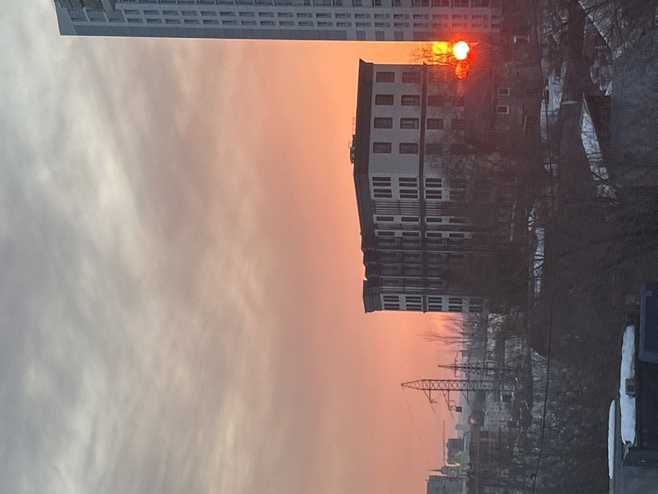 The image depicts an urban landscape in Perm, Russia, at either sunrise or sunset. The sky is stratified, transitioning from light grey with wispy clouds in the upper portion to a vibrant orange and deep red hue towards the horizon. A distinct, intensely red-orange solar disc is partially visible in the lower right quadrant, obscured by a tall, modern, light-colored high-rise building with multiple windows.
In the mid-ground, centered, is a five to six-story building with a flat dark roof and light-toned facades featuring repetitive dark vertical elements. Below this structure, an older, smaller, dark-colored building is partially visible. Bare deciduous trees, devoid of foliage, are distributed across the mid-ground and foreground, silhouetted against the bright sky and partially obscuring structures.
In the distant left, a cityscape is visible, including a large metallic lattice tower, likely an electricity pylon, and a domed structure topped with a cross, suggesting a church. Patches of snow are evident on ground surfaces and rooftops in the foreground, indicating cold weather conditions. No human figures or discernible activities are present.