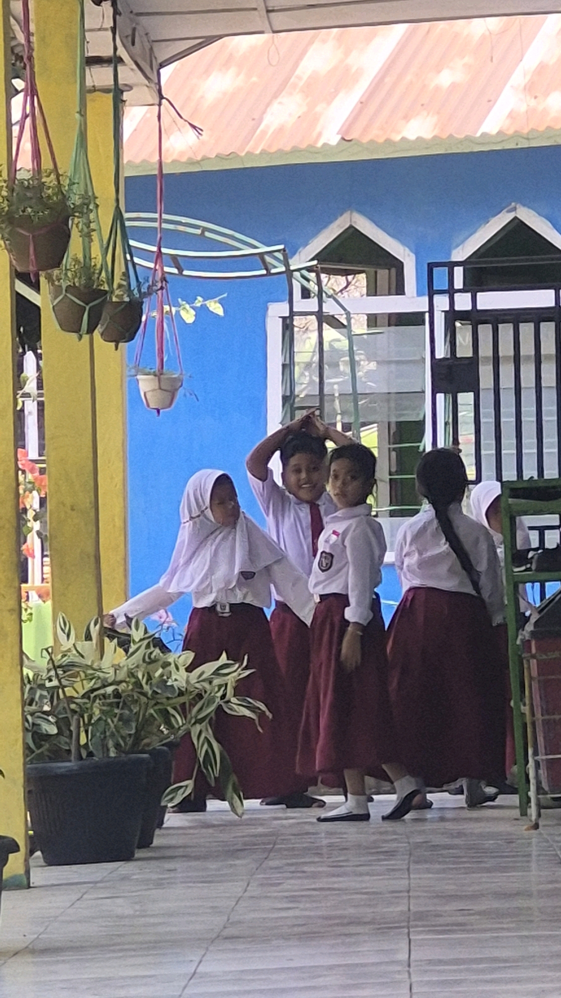 This image depicts a group of four young students, likely in uniform, standing on a tiled patio in front of a blue building with a red-tiled roof. The students are wearing white shirts and maroon skirts or pants. One student in the foreground is wearing a white hijab. Two students in the middle are smiling and looking towards the camera, with one of them playfully touching their head. Another student in the background is facing away from the camera. The environment includes hanging potted plants and a black metal fence. The scene appears to be outdoors, possibly at a school in Putak, Indonesia, given the architectural style and tropical vegetation.