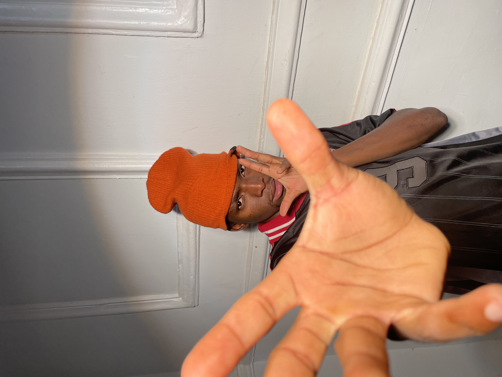 The image depicts a man wearing an orange beanie, a black and gray sports jersey with the number "23", and a red and white striped collar. The man is positioned indoors against a white paneled wall. He is looking directly at the camera. His right hand is raised in front of his face, partially obscuring his face. The background is out of focus. No details confirm the image's location in Barakin Akawo, Nigeria.