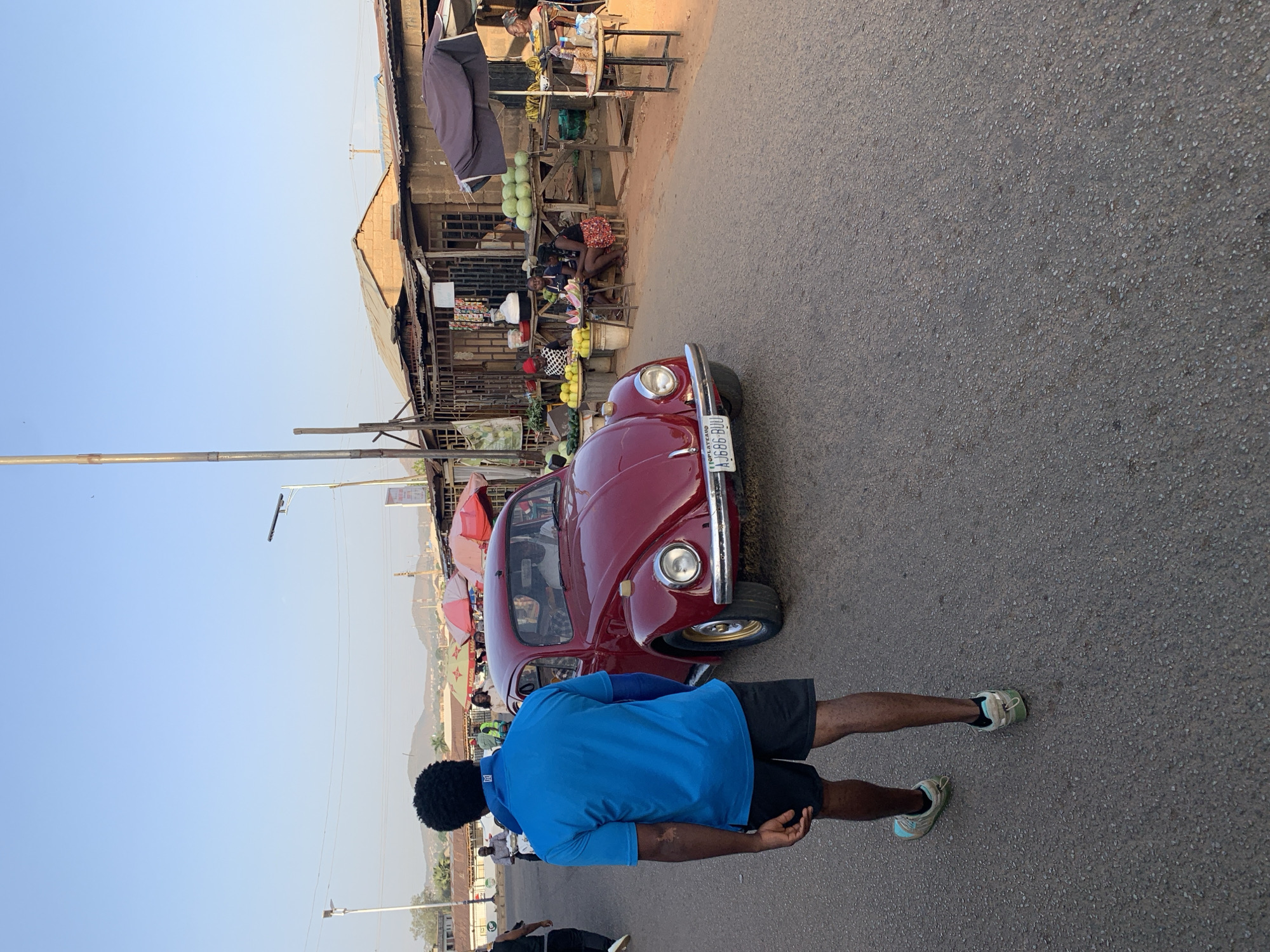 A street scene in New Stadium, Nigeria, shows a person standing on an asphalt road next to a red Volkswagen Beetle. The person is wearing a blue shirt, black shorts, and sneakers. Behind the car, there is a market with stalls selling various goods. People are present in the market area. The sky is clear and blue.