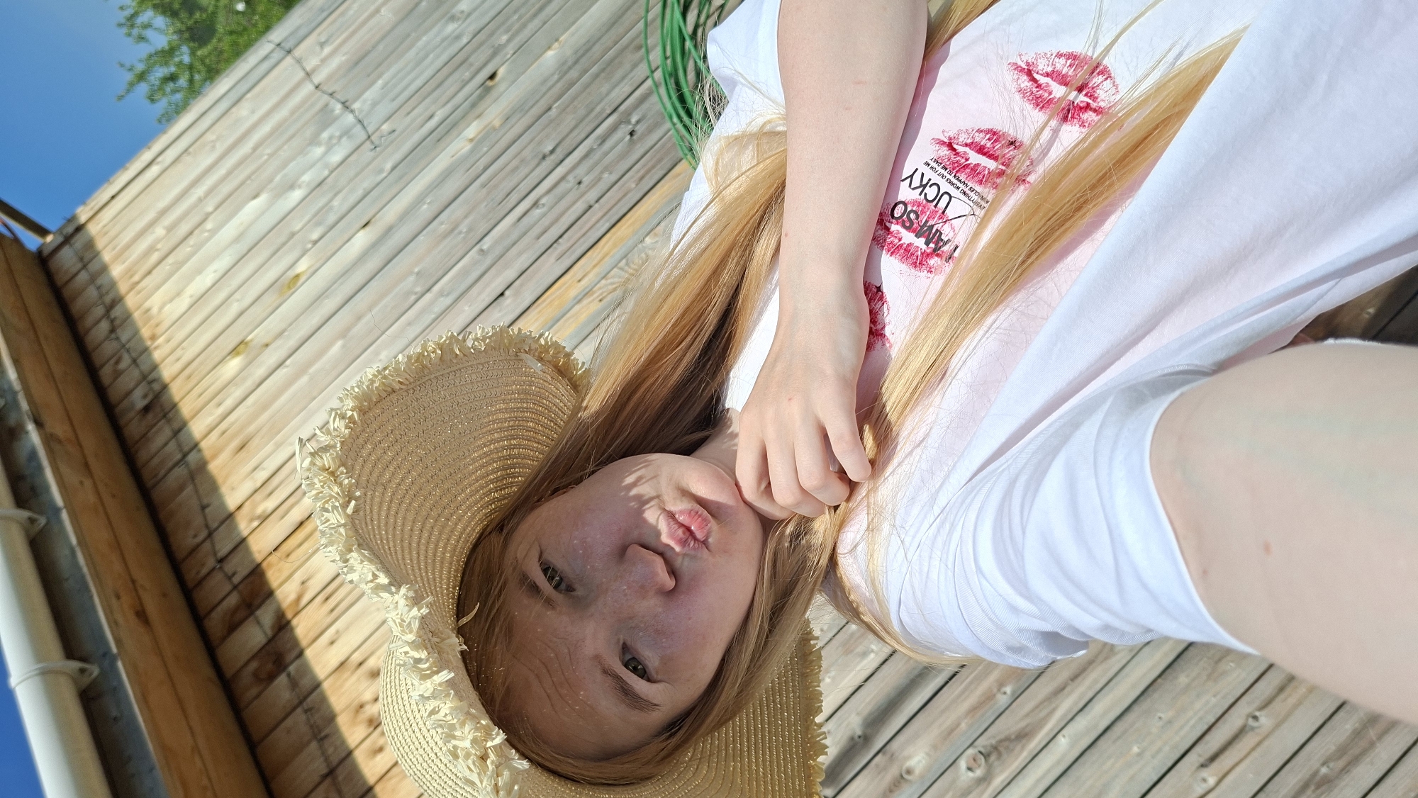 The image presents a young woman lying on what appears to be a wooden deck or platform. She is wearing a beige straw hat with a frayed edge and a white t-shirt over a pink undershirt with the words "I AM SO LUCKY" and red lipstick kiss marks printed on it. She is positioned at an angle, with her head close to the bottom-left corner of the frame and her body extending upwards. One hand is near her face. The background includes a wooden wall or structure to the left and potentially vegetation or sky visible in the top-left corner. Based on the provided location, the scene likely depicts a relaxing or leisurely moment on a deck or patio in Большой Букор, Russia.