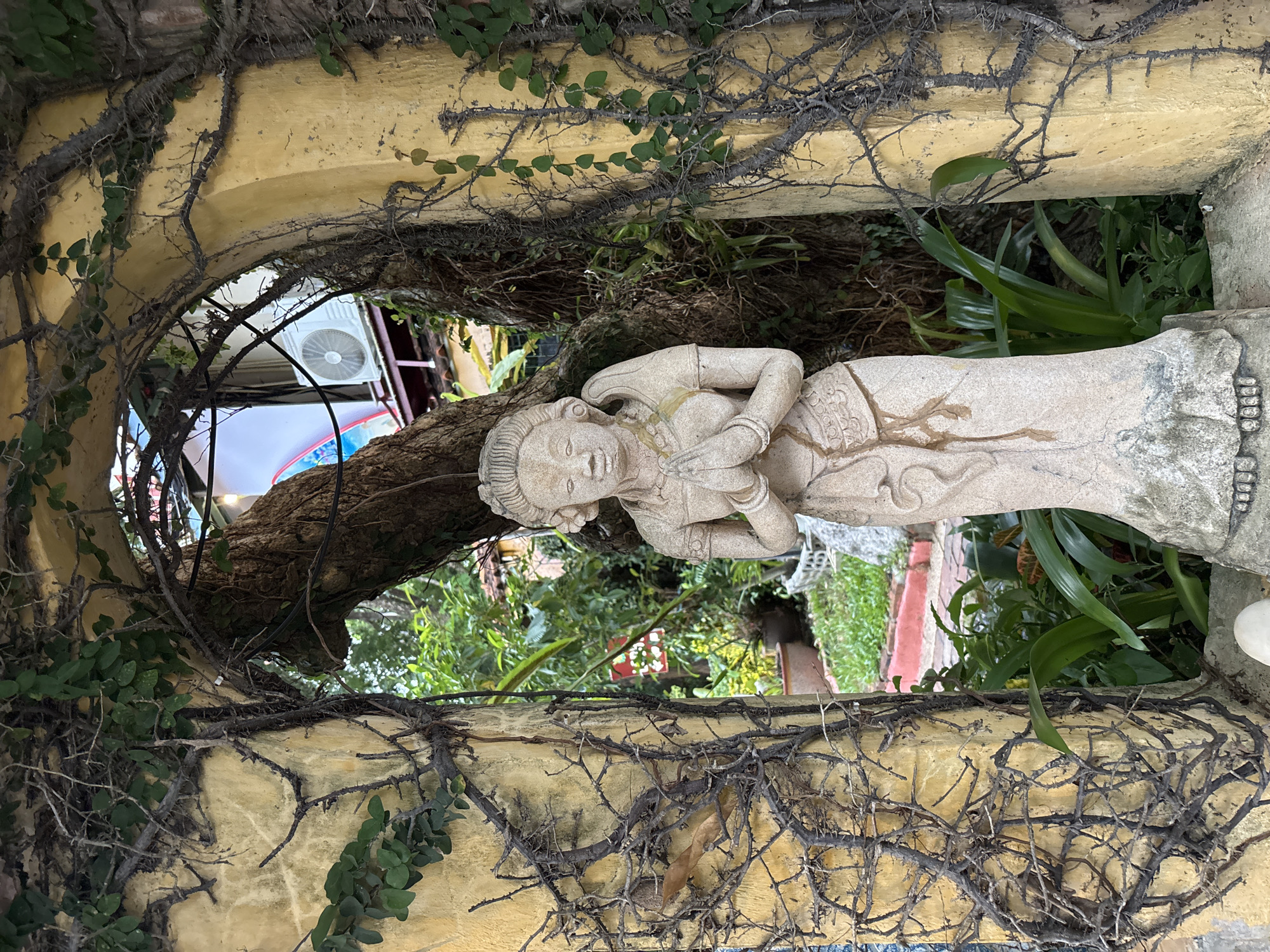 In the city of Chalong, Thailand, a stone statue of a woman with her hands clasped in a prayer-like gesture stands in front of an arched entryway covered in vines. The entryway is made of a textured, yellowish material. Behind the statue, lush green foliage is visible, along with parts of buildings and an air conditioning unit on a rooftop. The statue itself is detailed, depicting traditional clothing and hairstyle. The overall environment suggests an outdoor garden or courtyard setting.