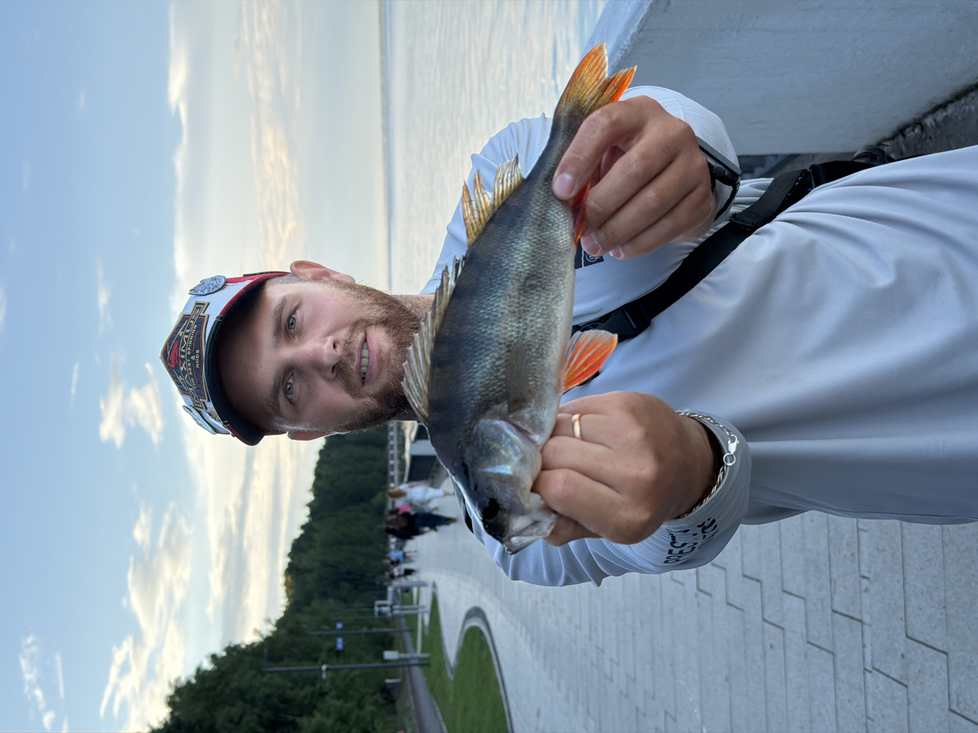 A man with a beard, wearing a white long-sleeved shirt and a baseball cap, stands on a paved walkway next to a body of water. He holds a fish in his hands, displaying it to the camera. The walkway is lined with a low white wall on the right, and on the left, there are trees and a path with people walking in the distance. The sky is cloudy and appears to be late afternoon or early evening. The location is implied to be Chaikovsky, Russia, based on the provided context.