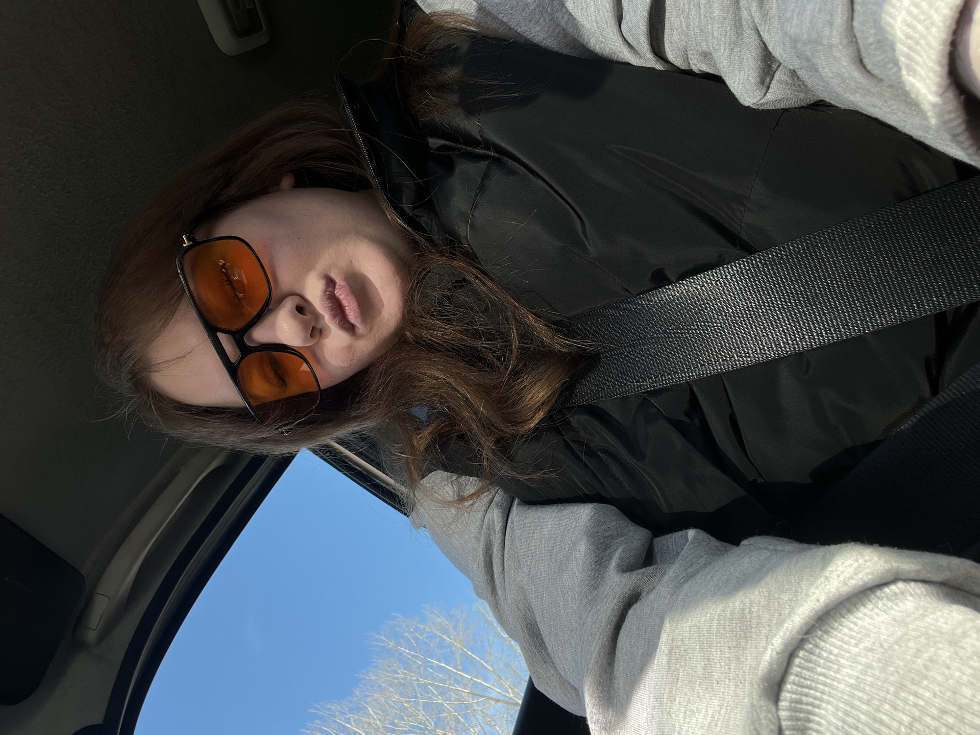 A single female individual is depicted in a selfie-style photograph taken inside a vehicle in Chaikovsky, Russia. She has light brown hair and is wearing dark-framed sunglasses with orange-tinted lenses. Her attire consists of a light grey long-sleeved top beneath a black sleeveless puffer vest. A black vehicle seatbelt is fastened diagonally across her torso. Her lips are pursed in a pouting expression. Through the vehicle's window, a clear blue sky and bare tree branches are visible, indicating an outdoor setting, likely during a season of dormancy for deciduous trees. No specific landmarks or precise locations within Chaikovsky are identifiable from the image.