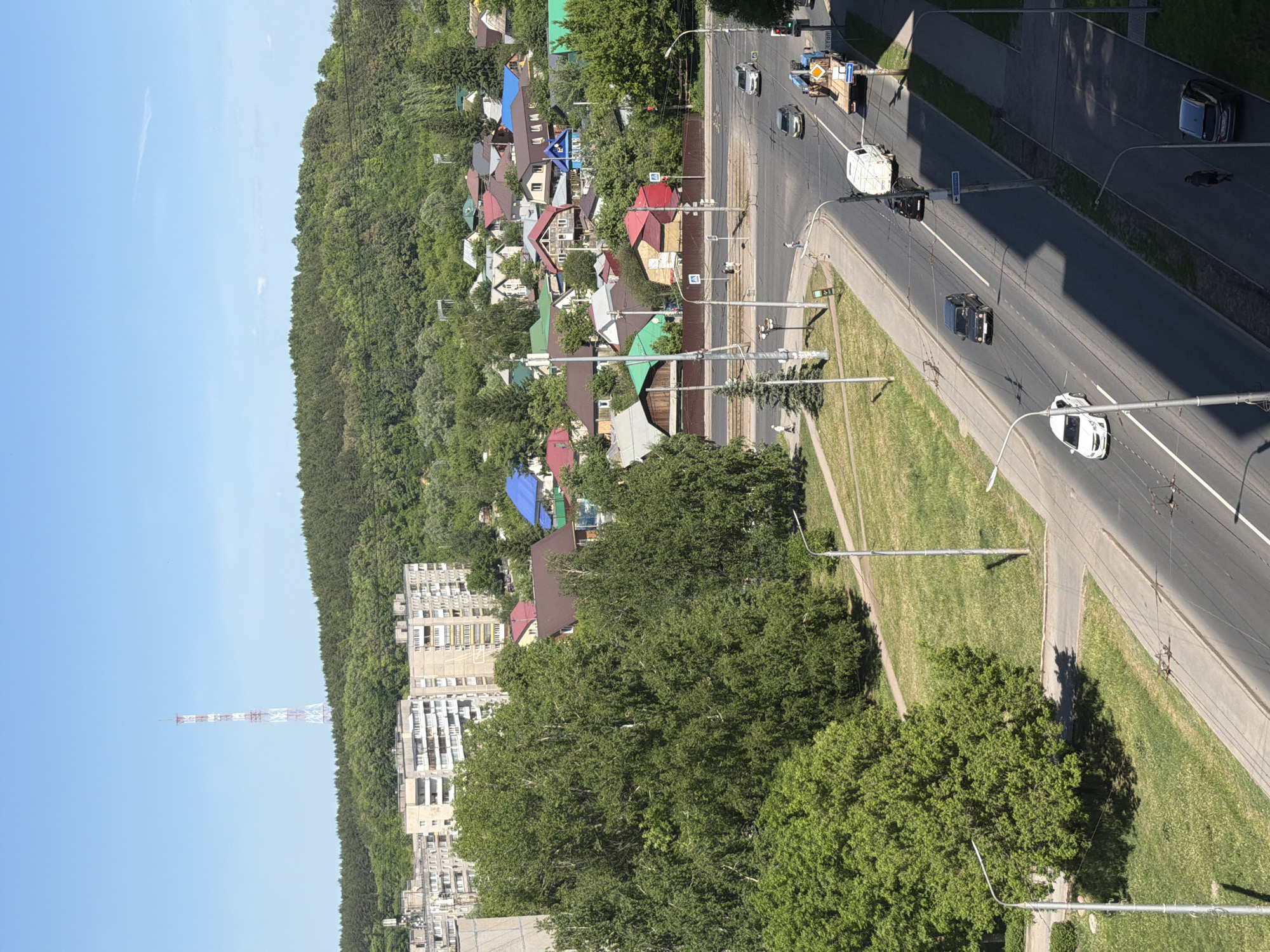 The image shows an elevated view of a cityscape. A multi-lane road dominates the right side of the image, with cars moving in both directions. The road is lined with streetlights and grass medians. To the left of the road is a mix of residential buildings, including individual houses with varied roof colors and taller apartment buildings, interspersed with dense greenery. In the distance on the left, a tall lattice tower can be seen against a clear blue sky. The image suggests a suburban or urban-fringe environment bordering a wooded area.