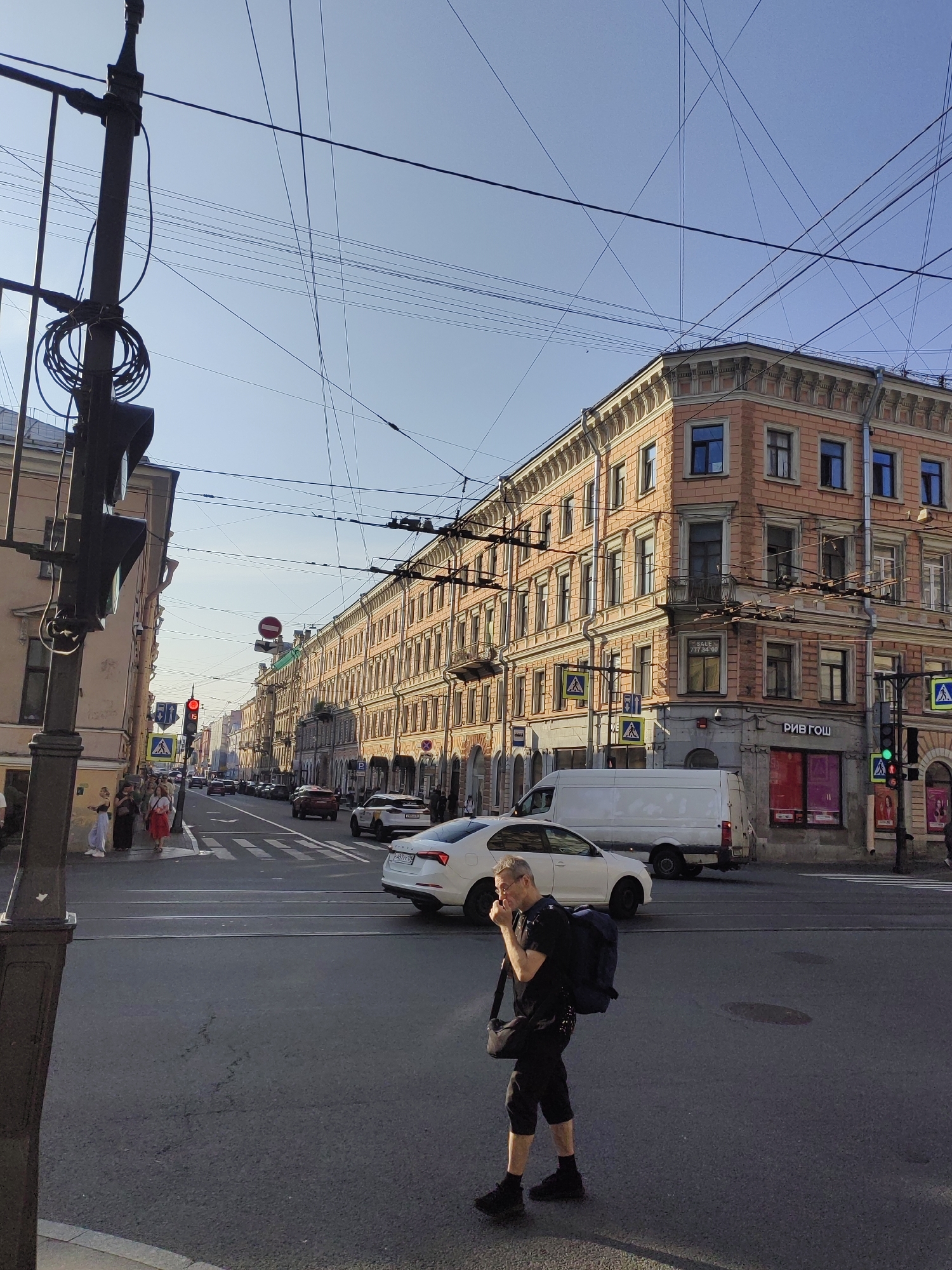 This media file captures a street scene in Saint Petersburg, Russia, under a clear blue sky, suggesting a pleasant, sunny day. The time of day appears to be late afternoon or early evening, with long shadows cast across the asphalt.
The main subject is a man walking across a crosswalk. He is wearing a dark t-shirt, dark shorts, black socks, and black sneakers. He carries a large dark backpack slung over one shoulder and a black messenger bag across his chest. He pauses briefly, bringing his hand to his mouth as if to sneeze or cough.
The scene unfolds at a busy intersection in front of a grand, multi-story building with salmon-pink brickwork. The architecture is typical of Saint Petersburg, featuring ornate cornices and numerous windows. Overhead, a complex network of trolleybus wires crisscrosses the sky.
Several vehicles are visible on the street, including a white van, a white car, and a red car. Traffic lights are present, with one showing red, indicating that traffic is stopped. Pedestrians can be seen on the sidewalks and at the crosswalk.
Visible text includes "РИВ ГОШ" in large Cyrillic letters on a storefront at street level on the pink building. Another sign with Cyrillic text is partially visible above the storefront. There are also street signs, including a "no entry" sign and pedestrian crossing signs with a yellow warning triangle. The overall impression is of a typical urban environment in Saint Petersburg during daylight hours.