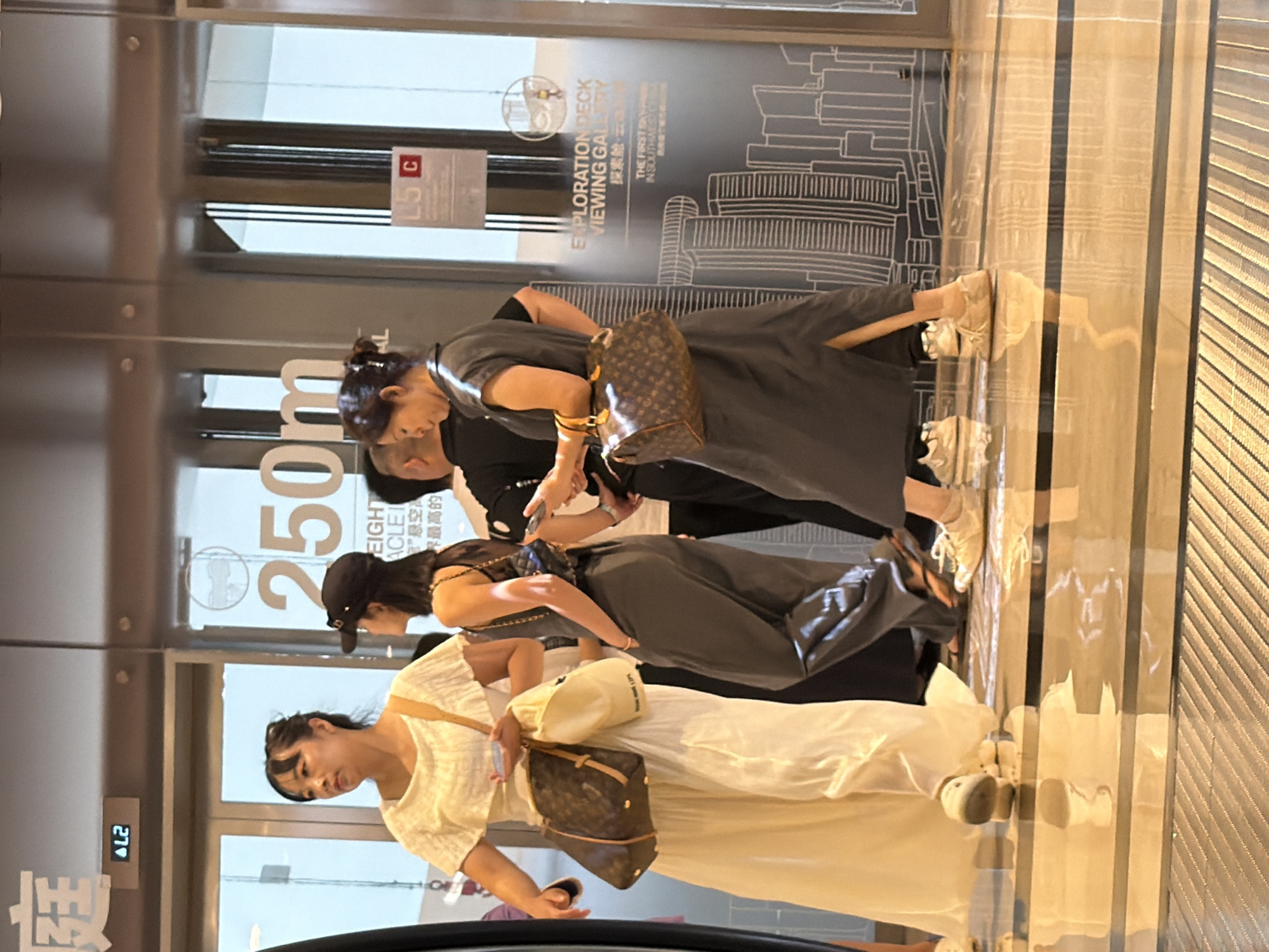 The image captures a scene inside a modern building, likely a shopping mall or entertainment venue, in Yuzhong District, China. Three women are the primary subjects. The woman on the left wears a long, flowing white dress and carries a brown and tan monogrammed shoulder bag. She is looking to her left. Next to her, a woman in a dark grey dress with a collared neckline is also carrying a similar monogrammed shoulder bag. She is looking down and slightly to her left, holding a mobile phone in her right hand. Behind them, partially visible, is another person in dark clothing.
The background features large glass doors and walls with graphics depicting city skylines. Prominently displayed on the glass is the text "250m" and "EXPLORATION DECK VIEWING GALLERY". A sign with "L5 C" is also visible. The flooring is highly polished and reflective, with dark horizontal lines. The overall environment suggests an indoor, public space with a focus on observation or entertainment.