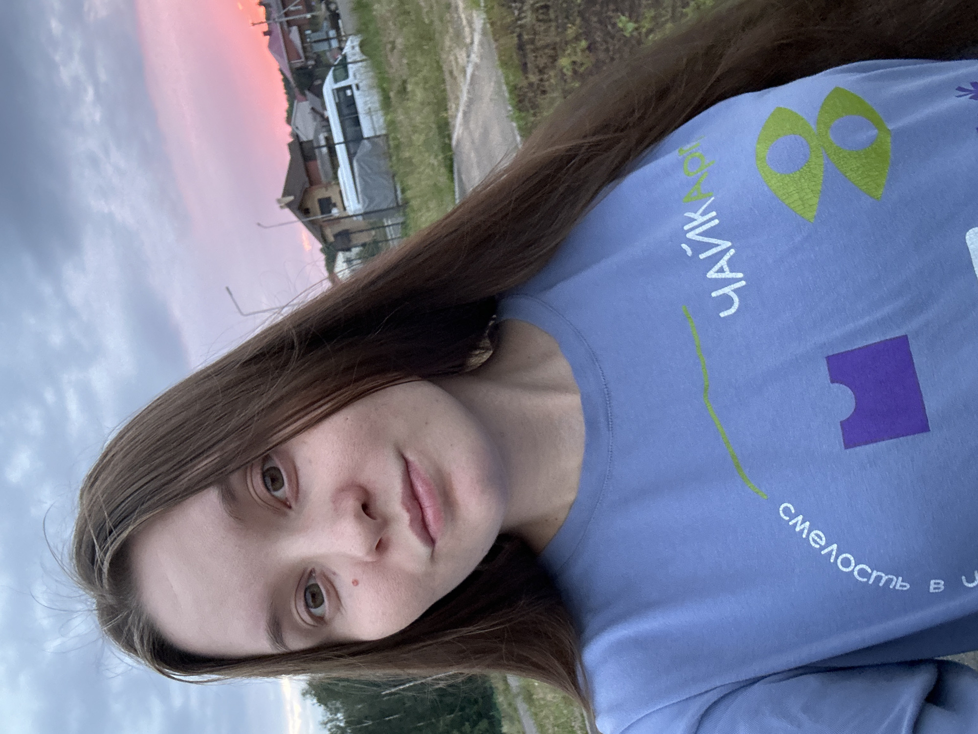 The image is a selfie taken outdoors. The subject is a young woman with long brown hair, looking directly at the camera. She is wearing a light blue t-shirt with a graphic design and text. The text appears to be in Russian and reads "ЧАЙКА АРТ" (Chayka Art) and "смелость в..." (bravery in...). The background shows a slightly cloudy sky with a pinkish hue, suggesting sunset or sunrise. There are houses with red roofs and a white van visible in the distance. The overall setting suggests a residential area, possibly a suburban or rural part of Noviy, Russia. The ground in the foreground is covered with grass and some paved areas.