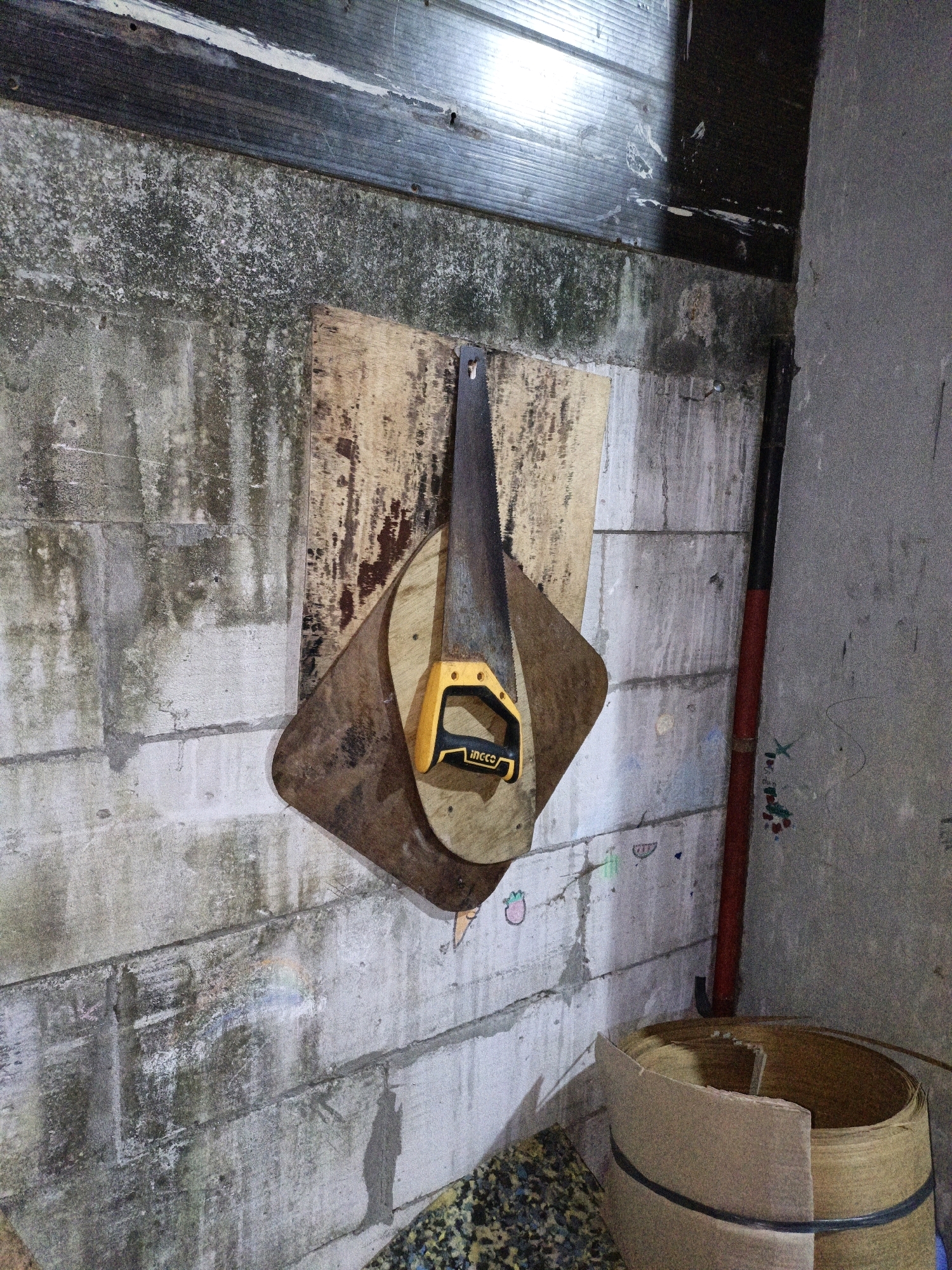 The image shows a saw with a yellow handle hanging on a wall. The wall appears to be constructed of concrete blocks and has some discoloration and moss. Above the concrete wall, there is a wooden planking. To the right of the saw, there is a red pipe and a grey concrete wall with some colorful markings. In the foreground, at the bottom right of the image, is a large roll of material, possibly cardboard or paper, held together by black bands. The overall scene suggests an indoor, possibly utilitarian space, like a workshop or storage area. The location in Cikal, Indonesia, is not visually evident from the provided image.