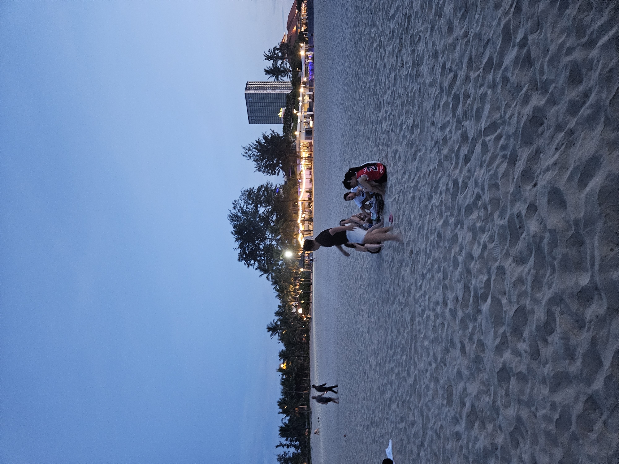 The image depicts a beach scene in Đà Nẵng, Vietnam, during the twilight hours. The sandy beach is populated with several individuals. In the foreground, a group of four women are seated on the sand, engaged in conversation. One woman wears a red basketball jersey with the number "2". To their left, a man in a black t-shirt and white shorts is walking away from the viewer. Further back, two men are walking in the distance.
Along the coastline, a row of palm trees lines the shore, with lights illuminating several structures behind them. These structures appear to be restaurants or resorts, with some of them featuring outdoor seating areas. In the background, a tall, modern building stands prominently against the darkening sky, suggesting an urban backdrop to the beach. The sky itself is a deep blue, indicative of dusk or early evening. The sand shows numerous footprints and slight undulations, common on a well-trafficked beach.
