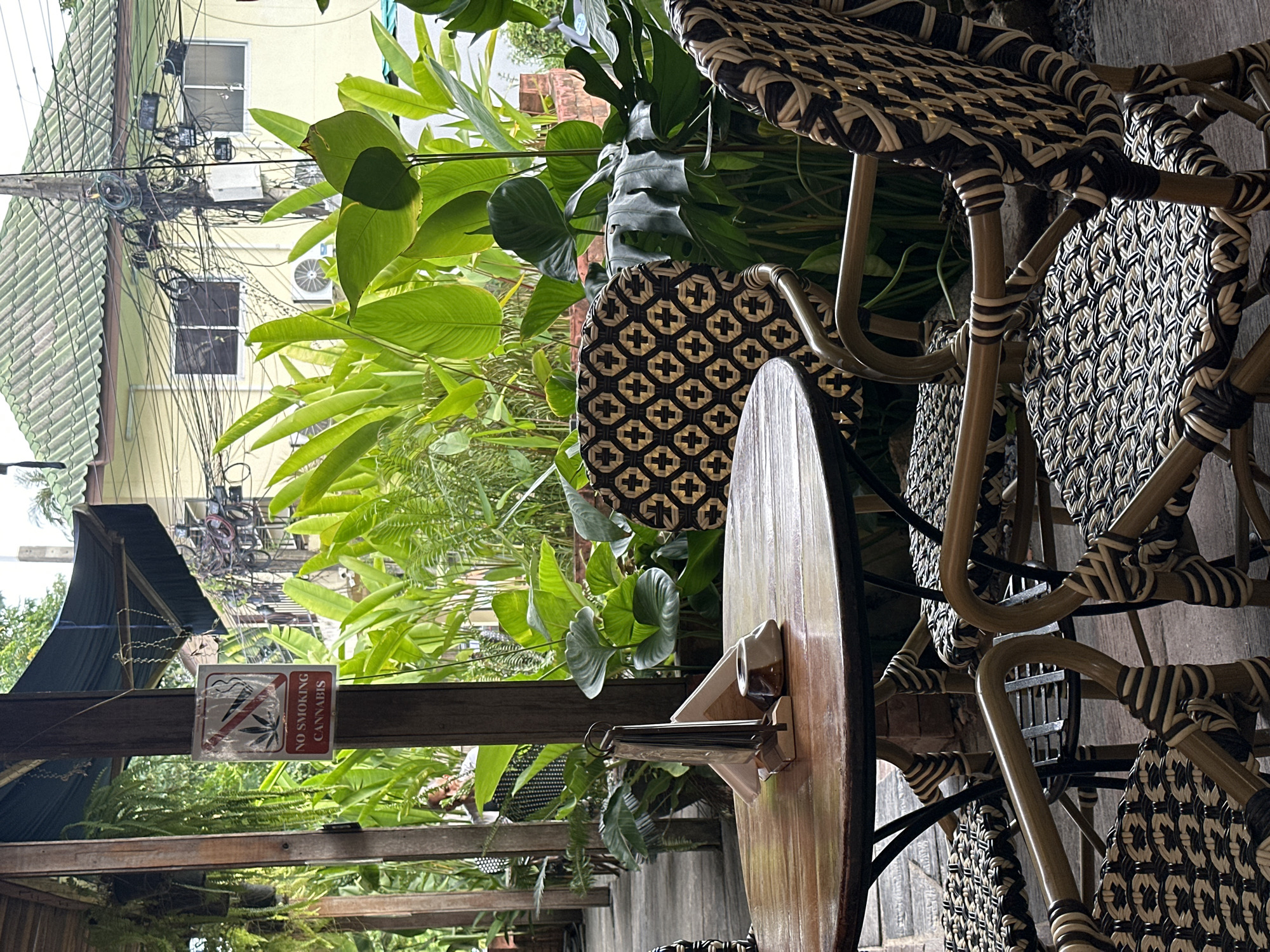 This image depicts an outdoor seating area at a cafe or restaurant on Ko Phangan, Thailand. A round wooden table with a metal base is set with a napkin holder and an ashtray. Three wicker chairs with a patterned design are arranged around the table. In the background, lush green foliage is visible, with a building and utility poles in the distance. A "No Smoking Cannabis" sign is affixed to a wooden post on the left side of the frame. The scene appears to be set during the daytime.