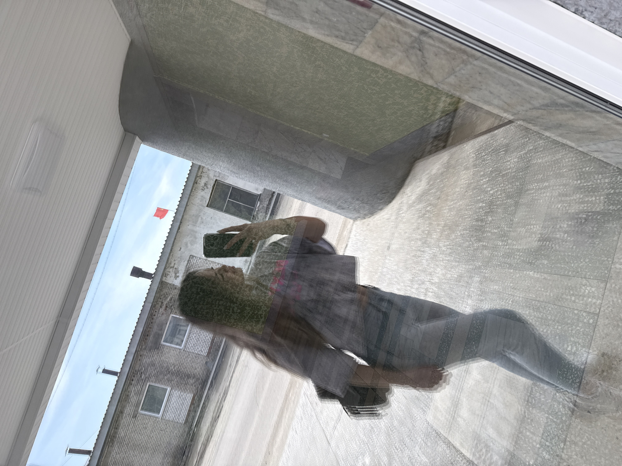 A woman is reflected in a window, seemingly taking a picture with her phone. Outside the window, there is a grey brick building with several windows and a red flag flying on a pole. There are also pipes extending from the roof of the building. The surrounding area appears to be an urban or industrial environment in the city of Unknown, Unknown.