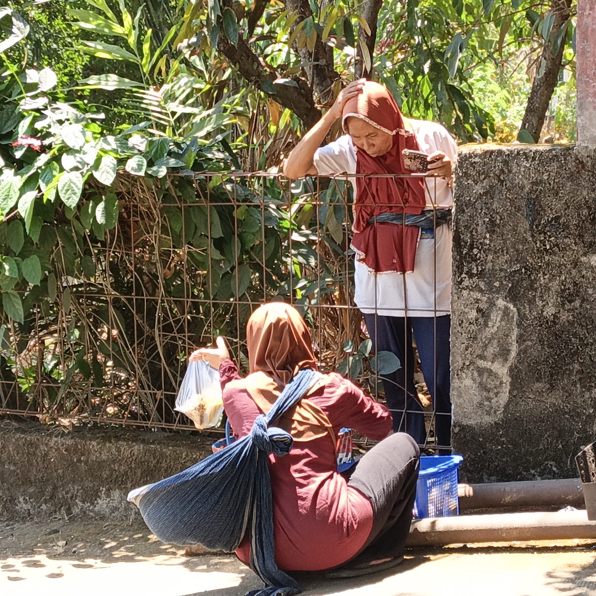 Two women are outdoors near a fence, possibly in the city of Mangli, Indonesia. The woman on the left is crouched down and holding a plastic bag. She is wearing a red top, a brown hijab, and dark pants. A blue cloth is tied around her body. The woman on the right is standing and leaning on the fence. She is wearing a red outer garment over a white shirt and dark pants, and a red hijab. She is holding a mobile phone and touching her head. The environment appears to be sunny with trees and foliage in the background. There is a concrete wall and a metal fence. A blue basket is visible near the standing woman.