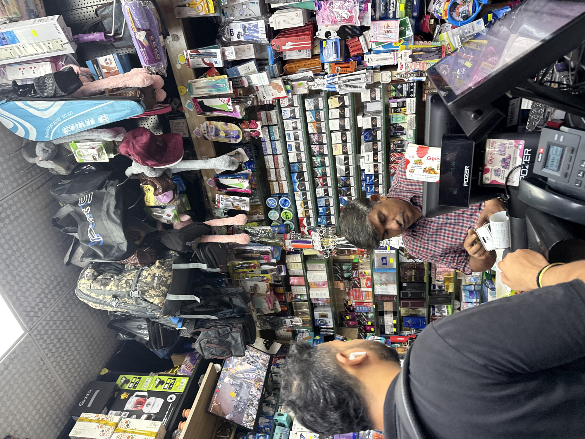 The image depicts the interior of a convenience store in Dubai, United Arab Emirates, with shelves stocked with various products. Two men are present. In the foreground, a man with dark hair and a beard, wearing a black t-shirt and white earbuds, is seen from behind, facing the interior of the store. To his right, a man with dark hair and a mustache, wearing a red and blue checkered shirt, is standing behind a counter with a point-of-sale system and a telephone. He is holding a receipt and appearing to be completing a transaction. The shelves behind them are filled with items such as backpacks, bags, electronics accessories, cosmetics, cigarettes, and other packaged goods. The lighting suggests an indoor environment, likely a small retail space.