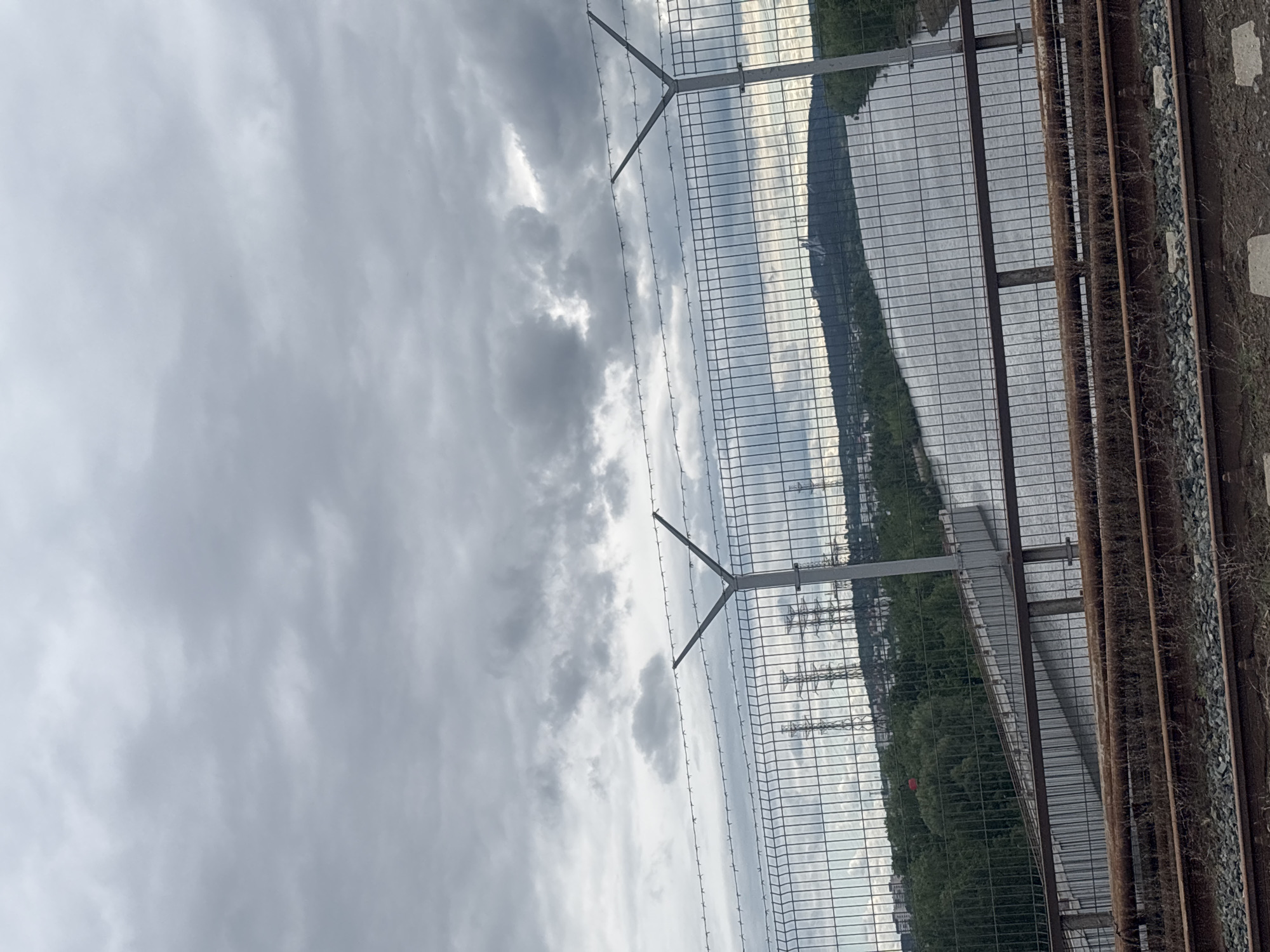 The image shows a railway track with a security fence alongside it in Chaikovsky, Russia. The sky is overcast with heavy clouds. On the right side of the image, the railway track is visible with gravel and railway sleepers. A metal security fence runs parallel to the track. In the background, there is a body of water and some vegetation. Utility poles can be seen in the distance.