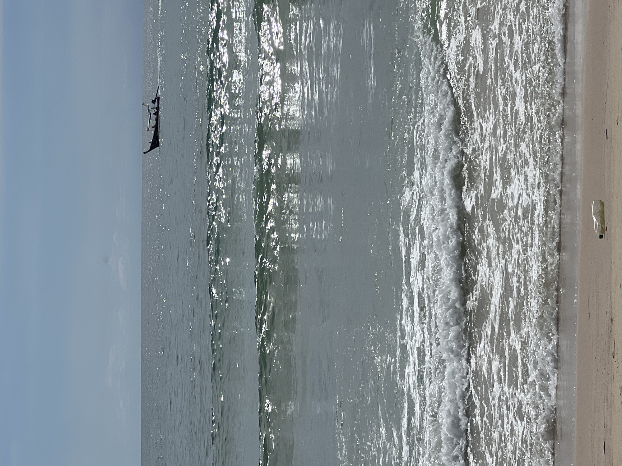 A lone, dark-colored boat with two people aboard sails on a calm, gray sea. The boat is a narrow, elongated vessel with a pointed bow and stern. One person is seated in the middle of the boat, and another person is standing at the stern, appearing to steer.
The sea is calm, with gentle waves lapping at the shore. The water reflects the bright sunlight, creating shimmering patterns on the surface. The sky is a pale, hazy blue, suggesting either a partly cloudy day or a hazy atmosphere.
The shoreline is visible on the right side of the image, with a sandy beach meeting the water. A single, white object, possibly a piece of plastic debris, lies on the sand near the water's edge. The overall mood of the image is peaceful and serene, with a sense of solitude.
The lighting suggests it is daytime, possibly late morning or early afternoon, given the brightness of the sun's reflection on the water. There are no clear indicators of a specific geographical location, other than it being a coastal environment. There is no visible text in the image.