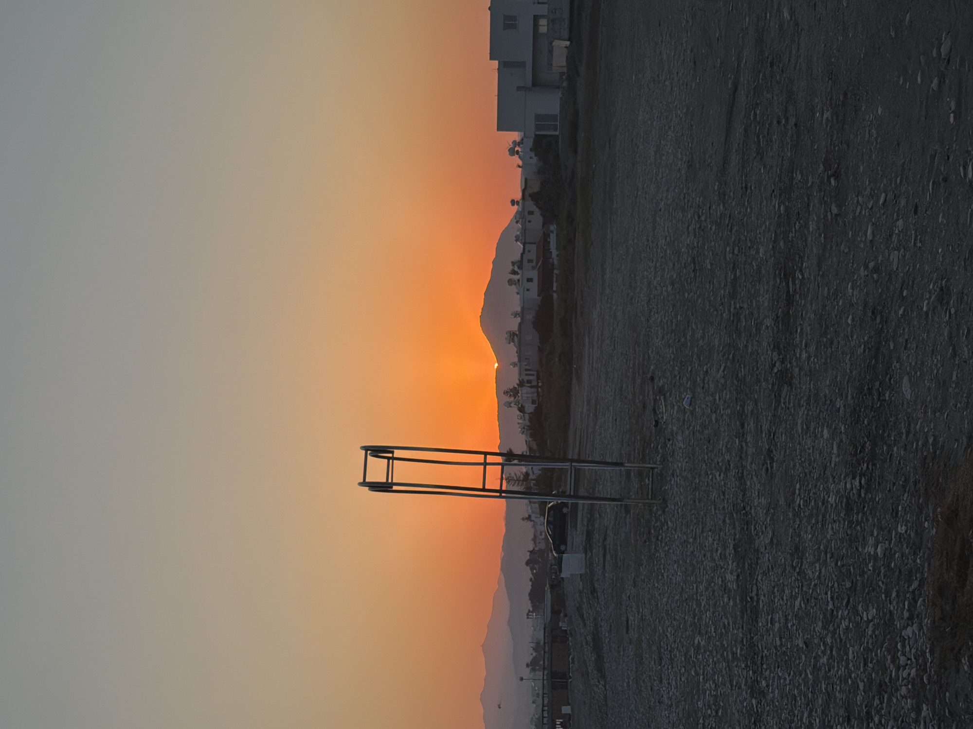 A tall, metal structure, likely part of a volleyball net or a similar sporting apparatus, stands on a gravelly expanse under a gradient sky of soft grey transitioning into a vibrant orange sunset. The sun, a bright orb, is partially obscured by a line of low, hazy mountains in the distance. The mountains are dotted with white buildings, suggesting a coastal town or resort. To the right of the frame, a dark, textured surface dominates, likely the ground of the beach or a similar area. The overall scene evokes a sense of calm and tranquility, characteristic of the end of a day by the sea. The time of day is clearly sunset, given the colors in the sky and the position of the sun. The location, Meneou, Cyprus, implies a Mediterranean setting, known for its beautiful sunsets and coastal landscapes. No text is visible in the image.
