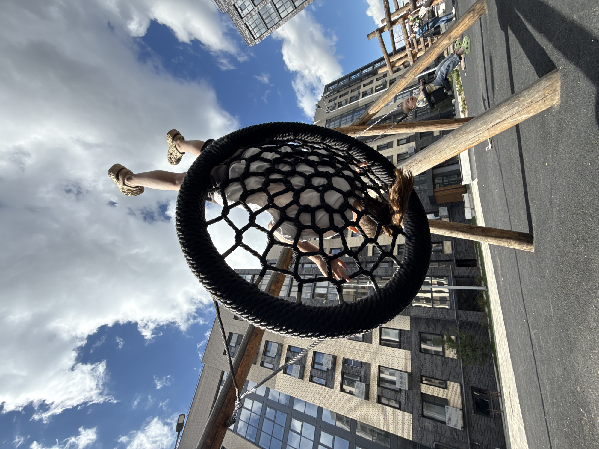 The photograph depicts a person swinging on a round net swing set in an urban environment. The view is from underneath the swing, looking up. The individual is positioned inside the swing, with their feet and legs visible towards the upper left of the frame. The net is black and has a circular shape with a thick rope border. Above the swing, there is a view of the sky with white, fluffy clouds and patches of blue. Tall buildings surround the swing set, visible through the netting. The buildings are made of brick and glass. The ground beneath the swing is paved with a dark surface. Wooden supports hold up the swing set and lead toward other play structures in the background. Another person is visible on a regular swing in the background.
