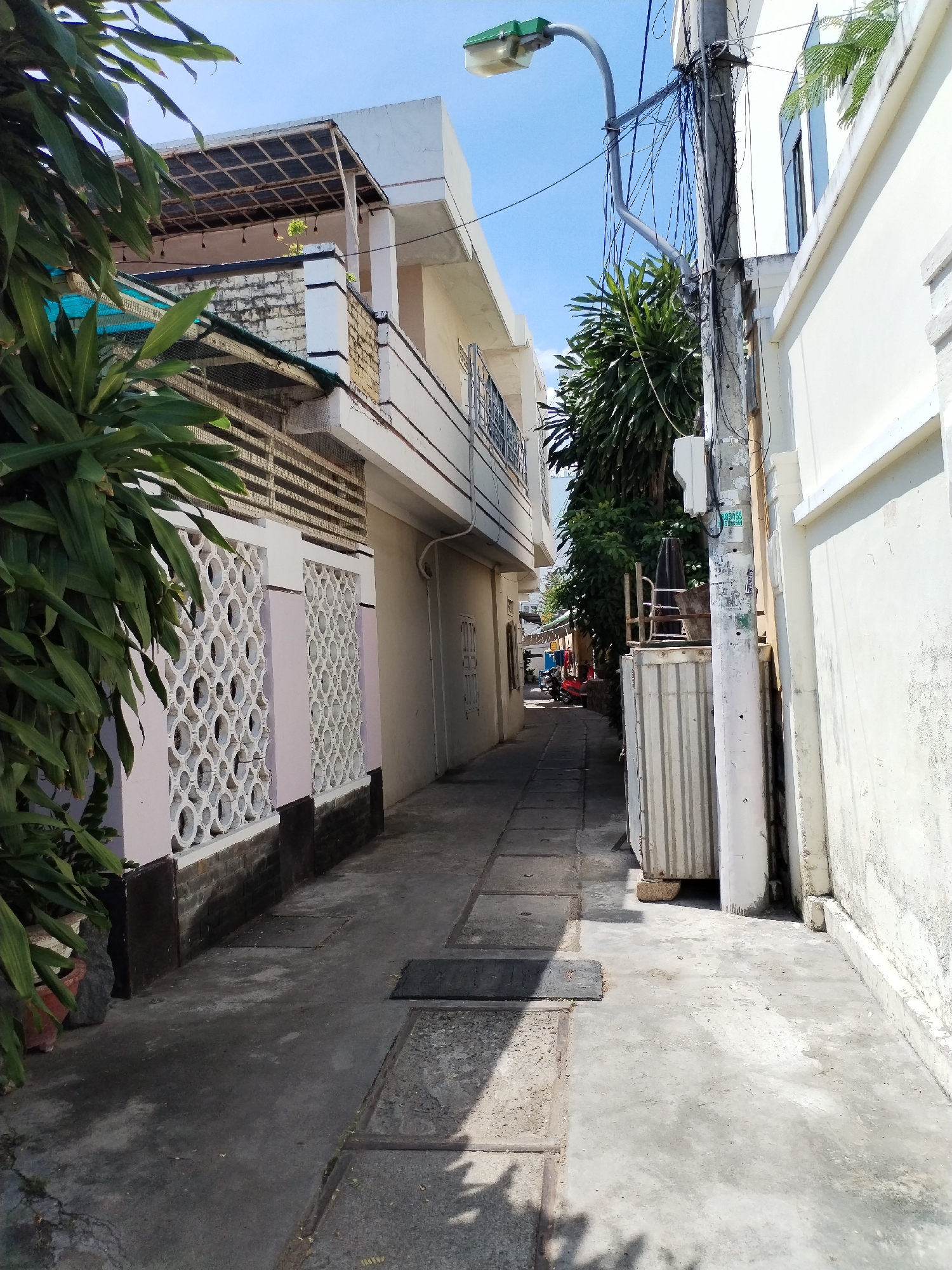 A narrow paved alleyway runs between two buildings with a clear blue sky above. On the left, a building features a patterned decorative wall with circular openings, painted pink and white, and a dark brick base. Lush green foliage from a large plant hangs over the wall. The building continues upwards with balconies and a simple roof structure. On the right, a white building with windows runs alongside the alley. A tall utility pole with numerous tangled wires stands prominently in the middle of the alley, a street lamp extends from it over the path. Further down the alley, a large grey metal transformer is visible next to the utility pole, with a smaller electrical box mounted on the pole. The ground is paved with concrete slabs, some of which are discolored or have visible seams. In the distance, at the end of the alley, a few individuals are visible, and a red scooter is parked. The scene appears to be a typical residential or commercial alleyway in a city.