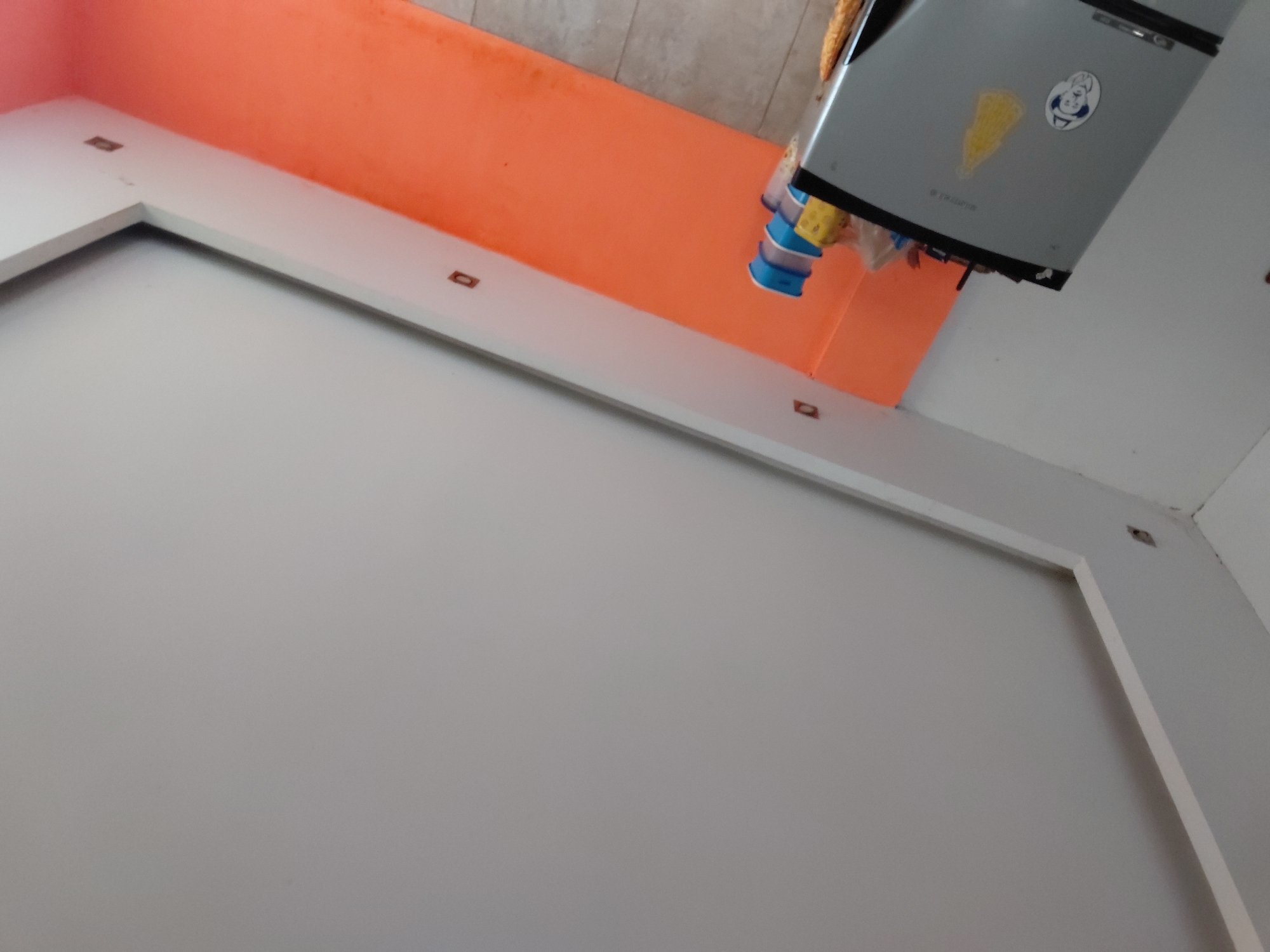 The image displays an interior domestic scene, captured from a low-angle perspective looking upwards and slightly tilted. The primary subjects are a gray top-mounted appliance, likely a small refrigerator or freezer, an orange wall, and white architectural trim.
The gray appliance is positioned high in the frame, featuring two prominent stickers on its front: one is a circular white sticker with a cartoon character's face, and the other is a yellow, wedge-shaped object, possibly depicting cheese or a fruit. Faint text, possibly "DURABA," is visible below the stickers. Several colorful plastic containers, including blue and yellow ones, are seen hanging from the underside of the appliance.
Behind the appliance is a vibrantly orange-painted wall, which shows some visible dust or discoloration along its upper edge where it meets what appears to be a ceiling or another wall. Below the orange wall and extending across the frame is a white L-shaped structure, which could be a shelf or part of a ceiling design. This white structure has small, rectangular, dark brown or bronze-colored inserts embedded at intervals along its top surface. The foreground is dominated by a plain light gray or off-white wall.
There is no discernible activity taking place, presenting a static view of a section of a room. The lighting suggests an indoor setting. The scene provides a glimpse into a residential space, possibly a kitchen or utility area, in Santa Ana, Venezuela.
