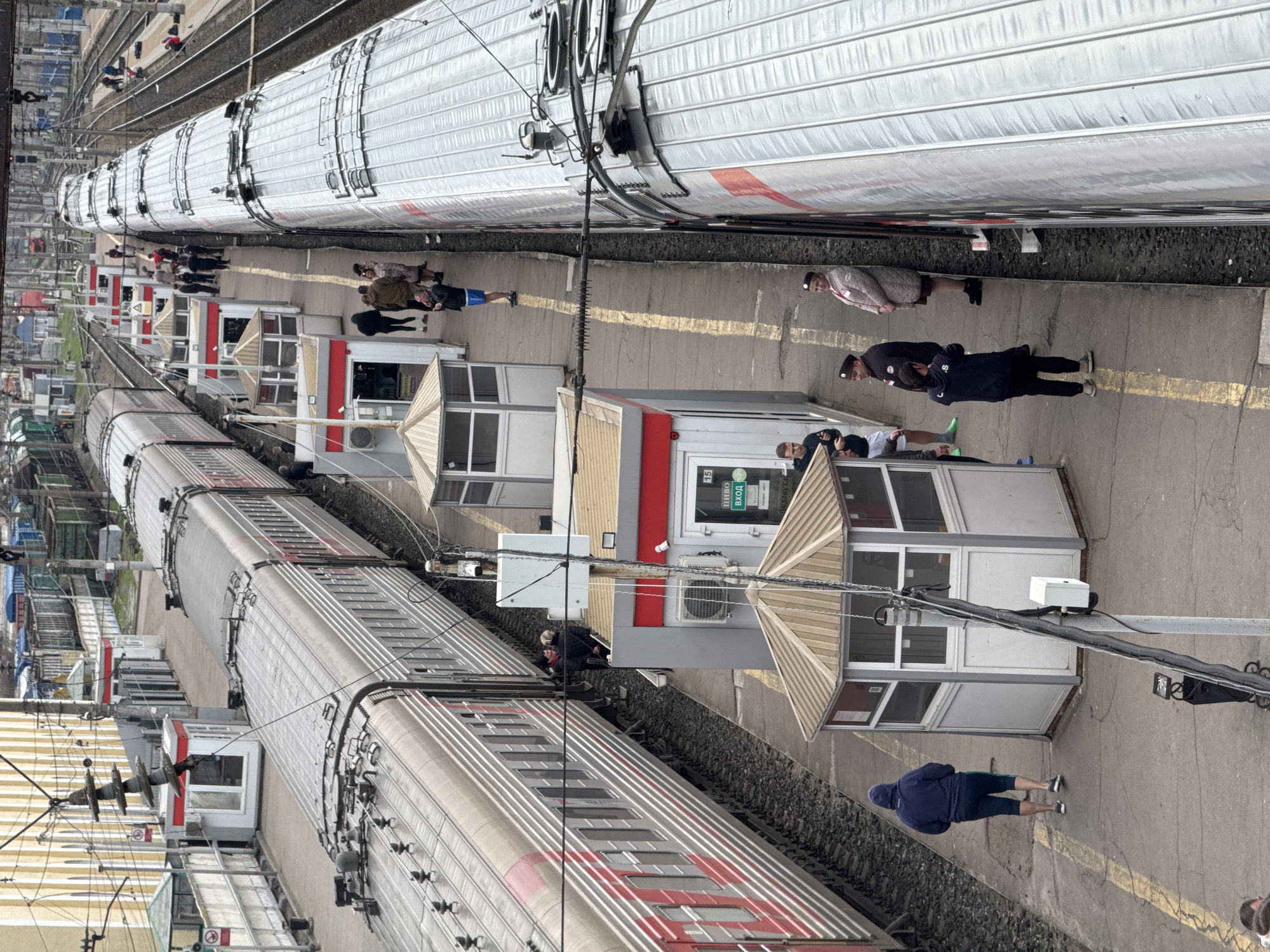 The image depicts a train station in Rossosh, Russia. Two trains are parked alongside a platform with small, booth-like structures. People are standing and walking on the platform between the trains and the booths. Overhead power lines are visible. The buildings are painted white and gray with red accents. The trains appear to be silver with red stripes. The platform is concrete.