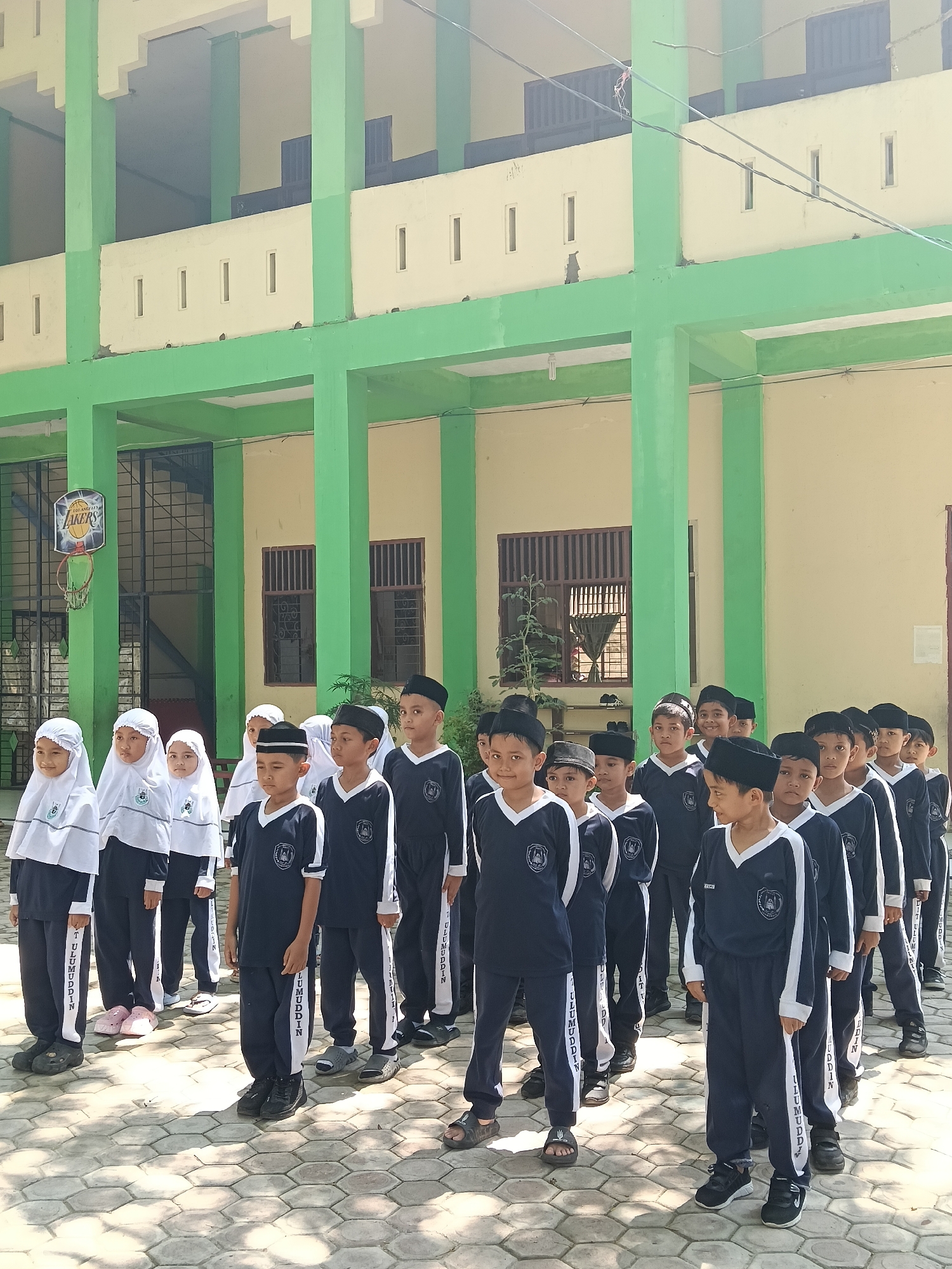 A group of young children, primarily boys with some girls, are depicted standing in disciplined lines outdoors. The children are dressed in uniforms; the girls wear white hijabs with long-sleeved tops and dark trousers, while the boys wear dark blue or black long-sleeved V-neck shirts and matching pants, many with black peci (kufi caps). The uniforms feature a white vertical stripe on the pant legs and sleeves, displaying the text "ULUMUDDIN."
They are standing on a paved area with hexagonal interlocking blocks, beside a multi-story building. The building features a distinctive design with prominent green pillars and beams, contrasting with cream-colored walls and railings. Open corridors are visible on the upper levels, and some windows are barred. On the left side of the image, a basketball hoop with a "LAKERS" logo on its backboard is visible, suggesting a school or recreational area.
The bright sunlight and pronounced shadows indicate it is daytime, likely mid-morning or early afternoon, on a clear, sunny day. The children's expressions are mostly attentive, suggesting they are participating in an assembly, a class formation, or a similar structured gathering. The attire, particularly the hijabs and peci, points to an Islamic educational setting, consistent with the specified location of Lhokseumawe, Indonesia.